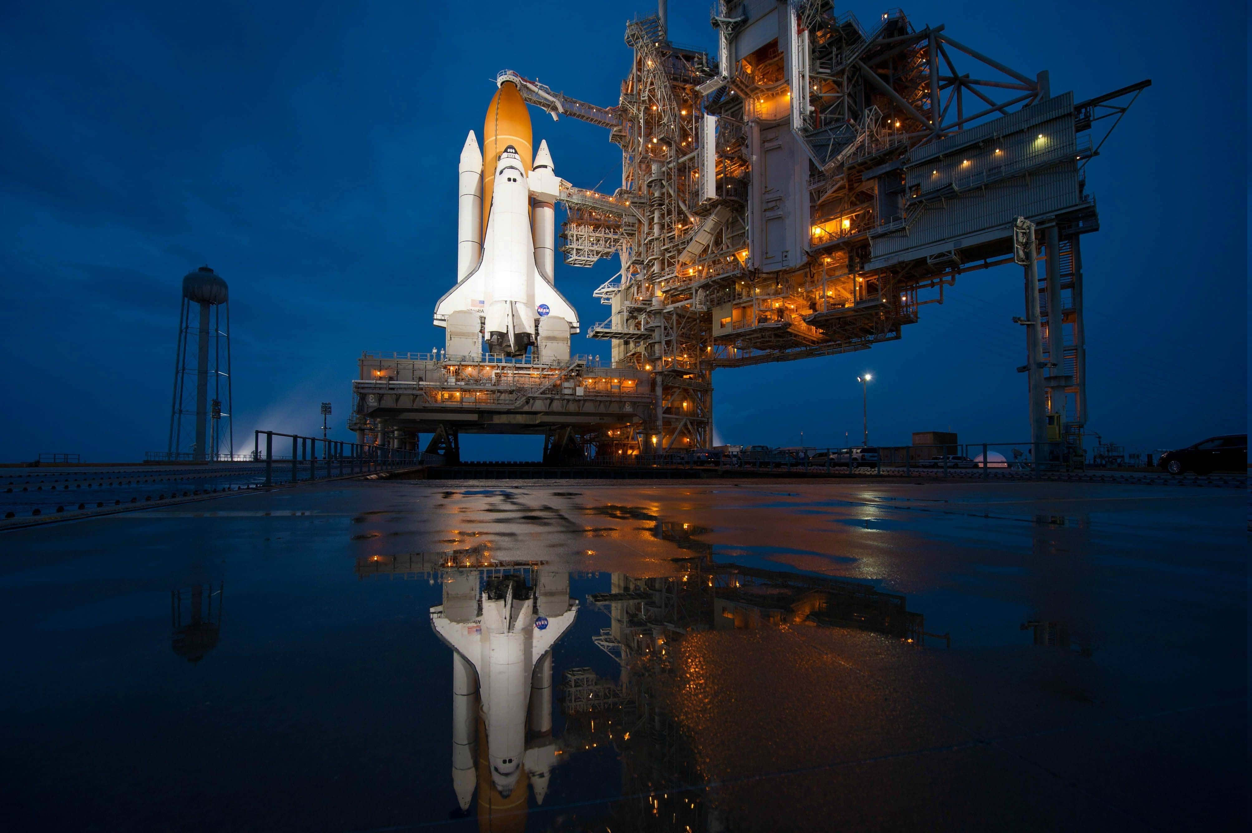 space shuttle 桌布 壁紙 — 一架太空梭在黃昏時分被照亮，矗立在濕漉漉的發射台上，其倒影在下面的水坑中閃爍。