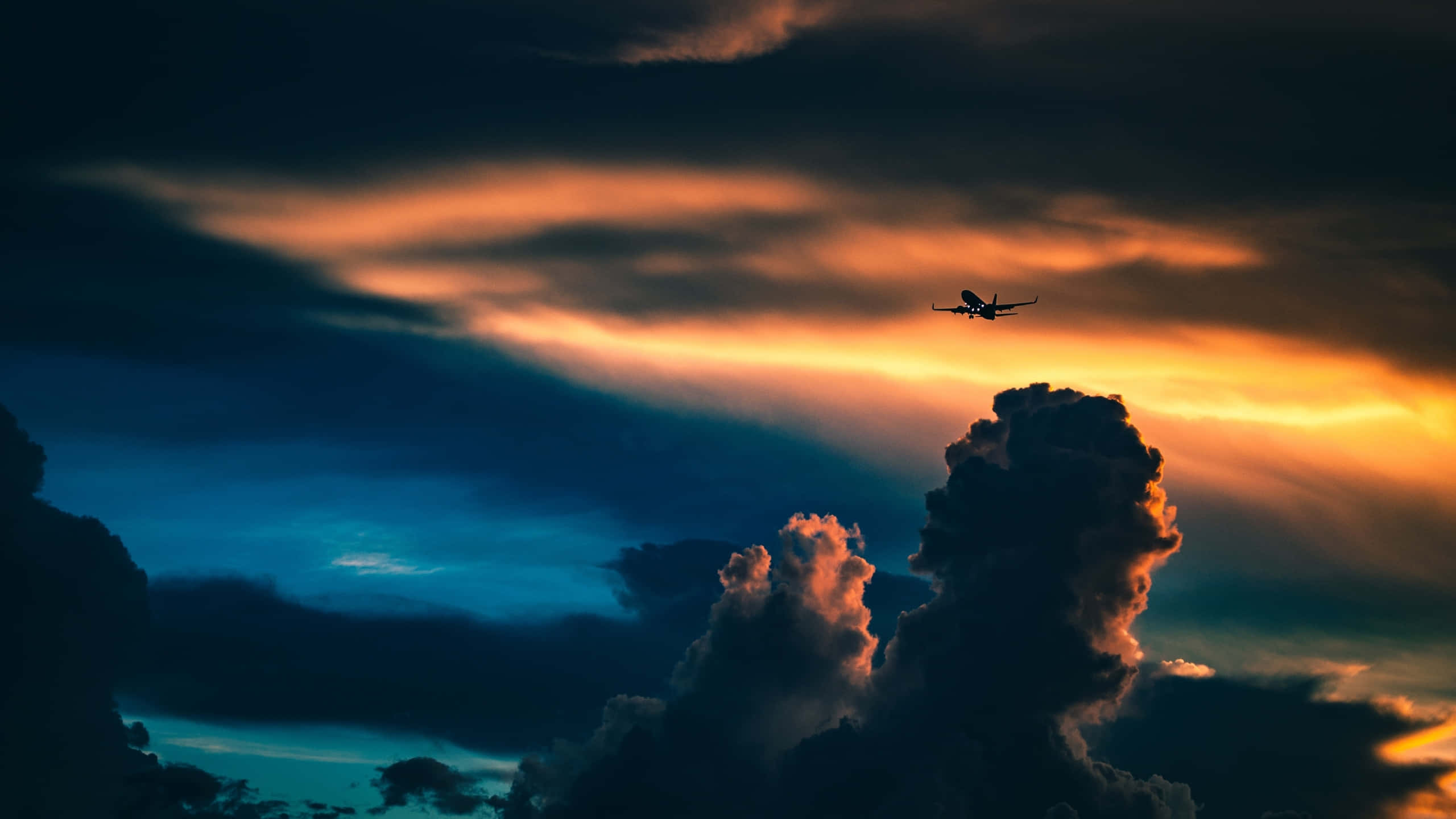 1440p Plane 桌布 壁紙 — 一架孤獨的飛機在黃昏時分，以火橙色和深藍色雲彩繪成的戲劇性天空為背景，呈現剪影。