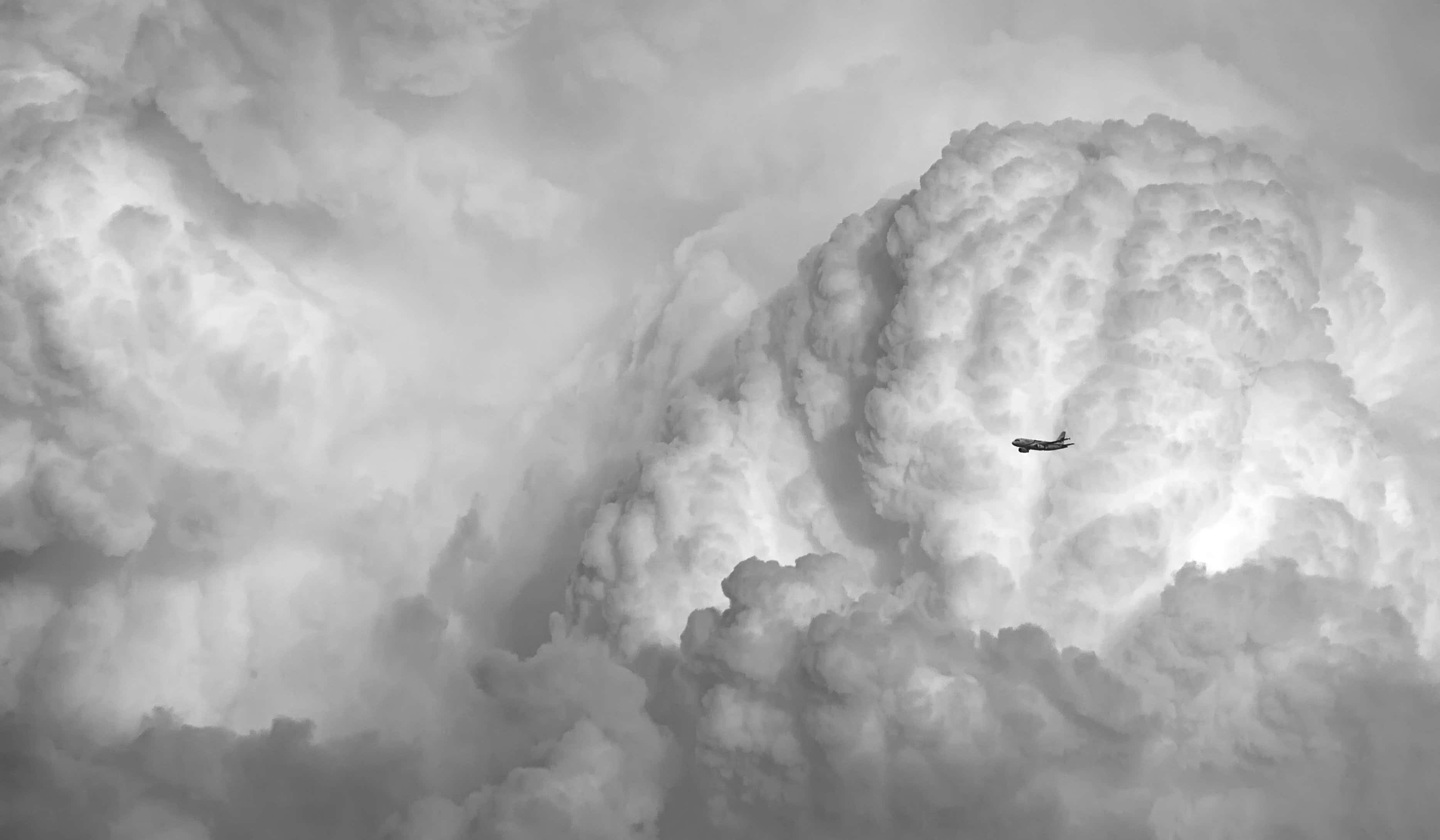 Black And White Storm 桌布 壁紙 — 一架孤獨的飛機飛過廣闊的單色天空，天空充滿了高聳、有紋理的雲層。