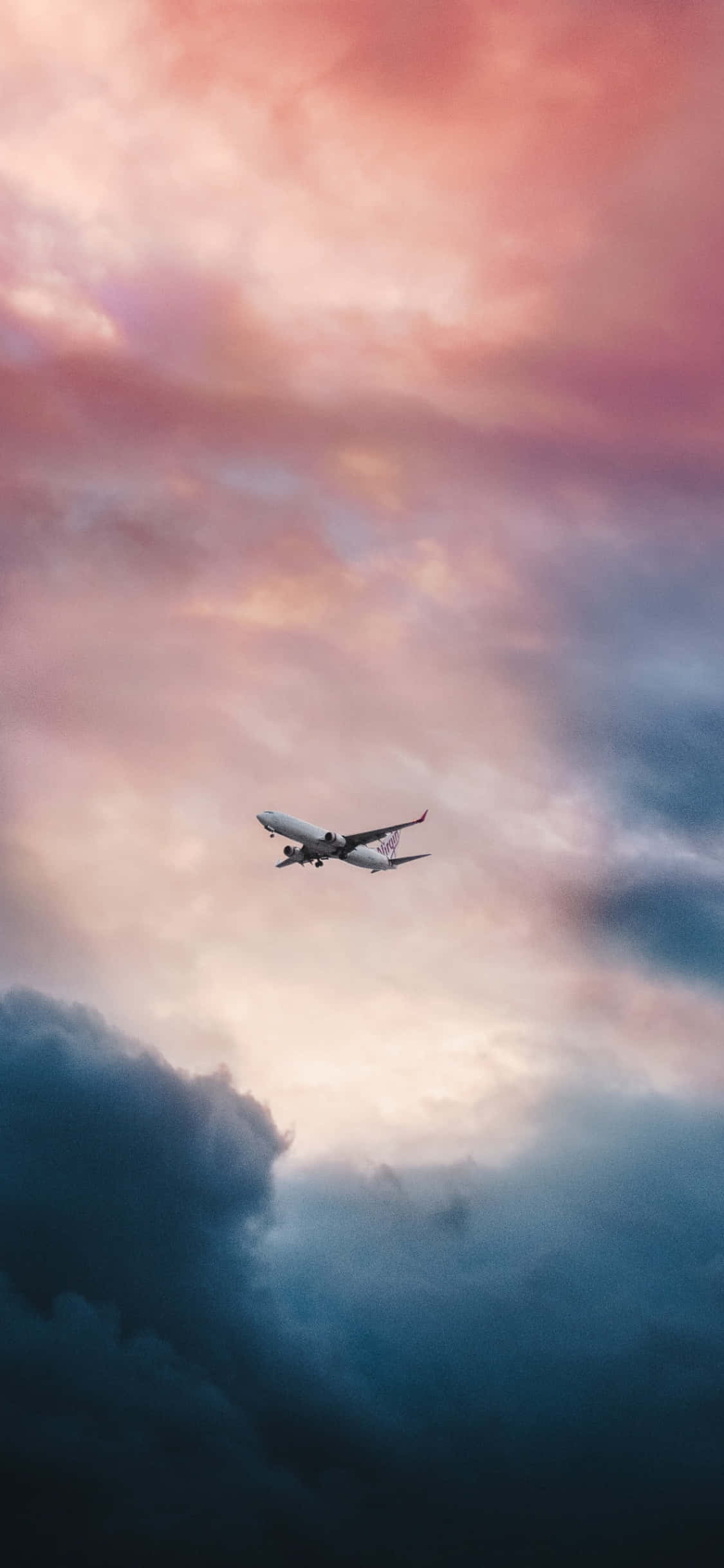 iPhone Xs Max Small Planes 桌布 壁紙 — 一架客機在暮色中翱翔，穿過由粉紅、橙色和深藍色雲彩繪成的戲劇性天空。