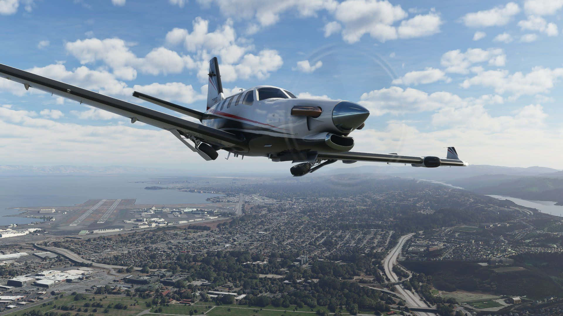 1920x1080 Microsoft Flight Simulator 桌布 壁紙 — 一架小型白色螺旋槳飛機在部分多雲的天空下低飛過一個有道路和水的沿海城市。