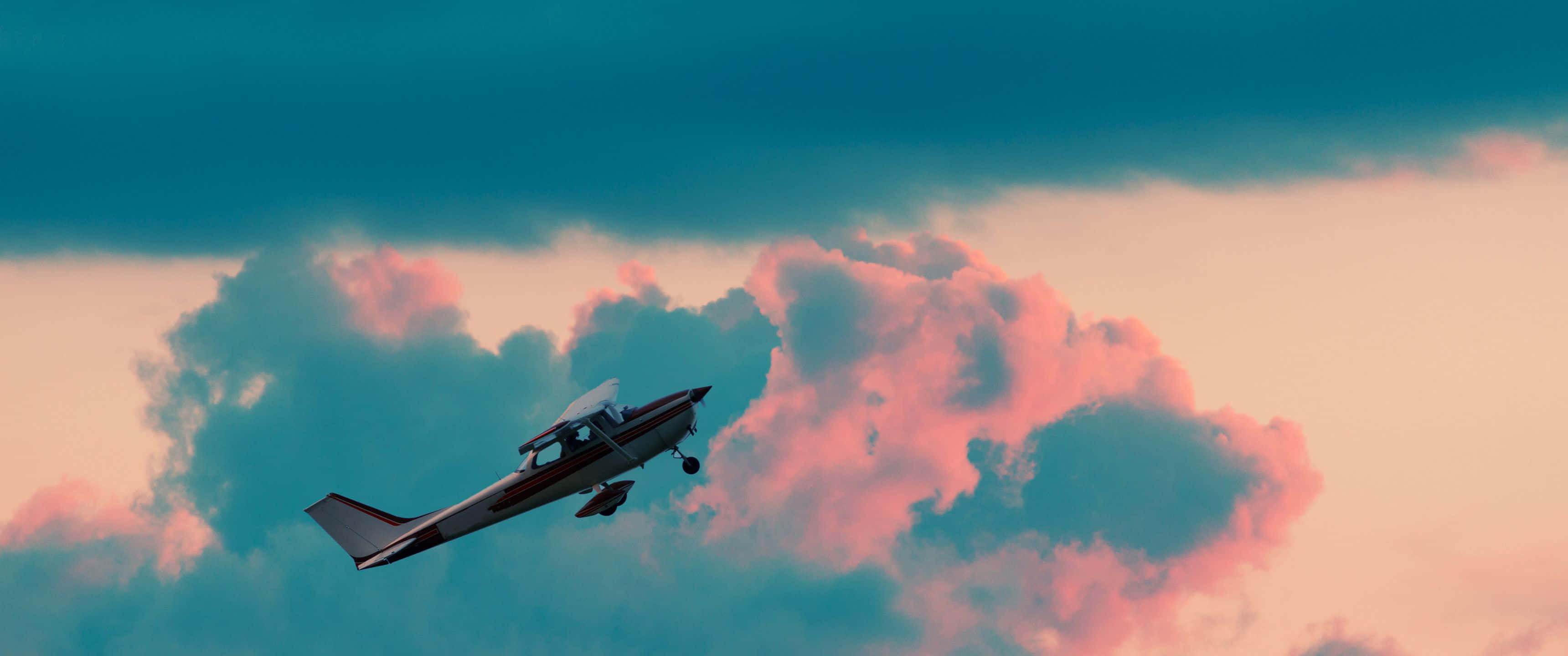 3440x1440p Small Planes 桌布 壁紙 — 一架小型飛機在壯麗的日落中，翱翔於充滿活力的粉紅色和青綠色雲層之間。