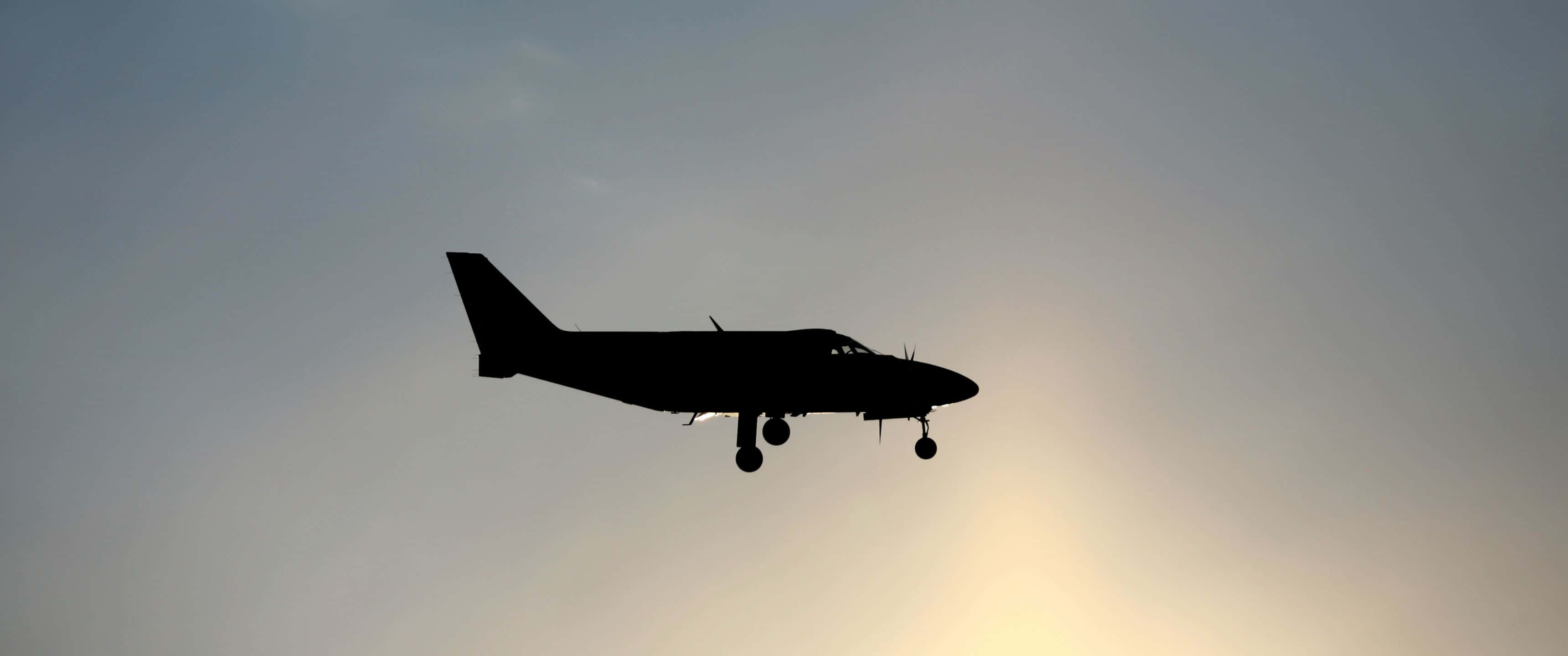 3440x1440p Small Planes 桌布 壁紙 — 一架小型飛機在日落時分，以柔和漸變的天空為背景，呈現剪影，起落架已放下。