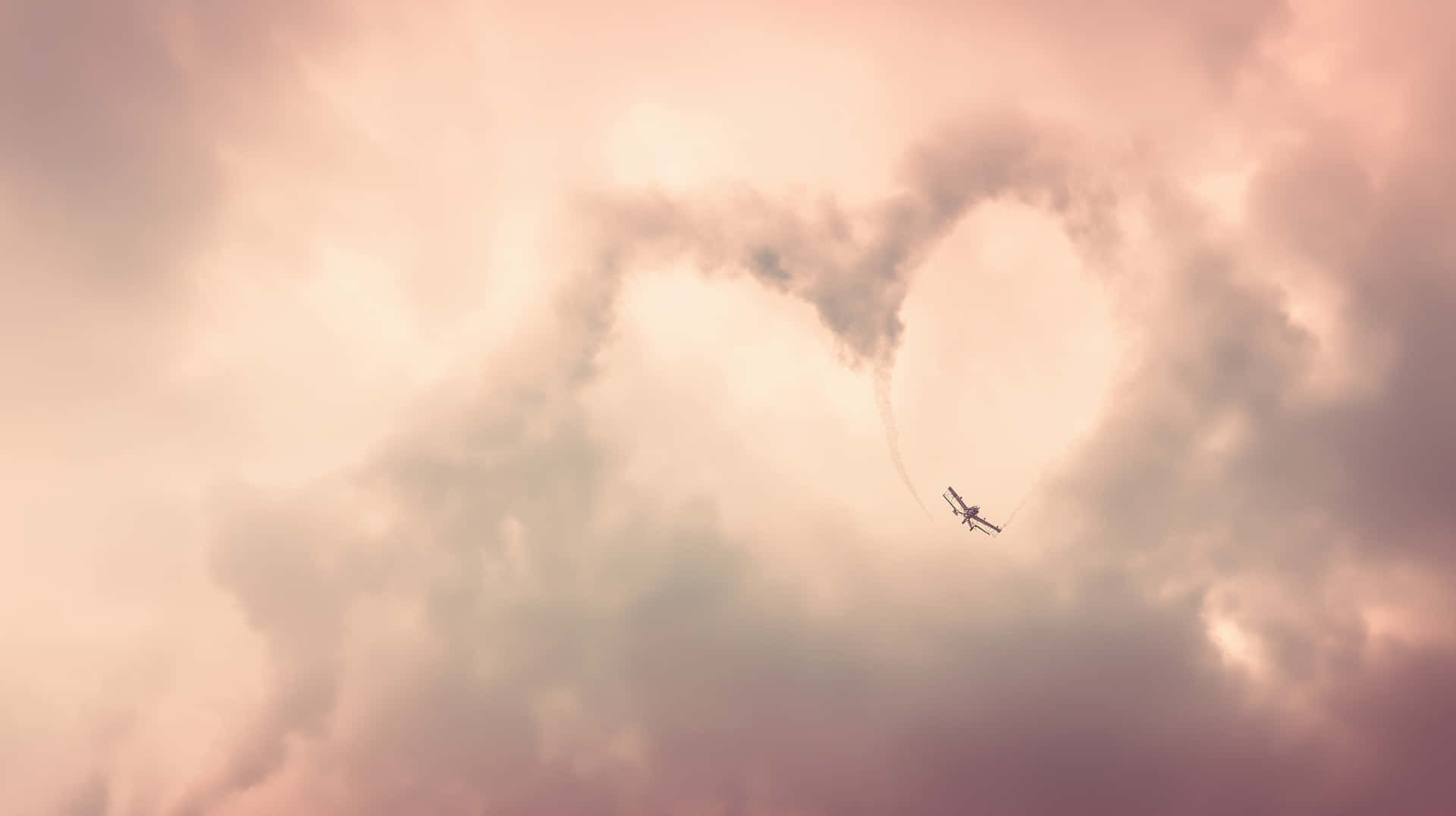 Biplane Creating Heart-Shaped Skywriting in Clouds 壁紙 — 一架小型飛機在柔和粉紅和蜜桃色的多雲天空中留下心形煙霧。