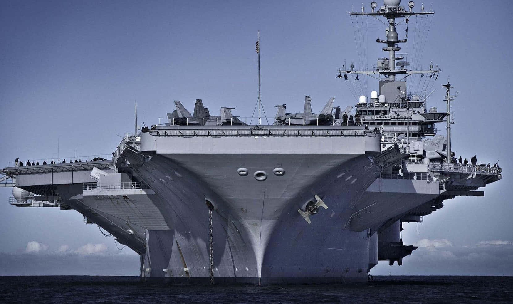 Navy 桌布 壁紙 — 一架巨大的灰色航空母艦佔據畫面，其傾斜的飛行甲板載有戰鬥機，背景是清澈的藍天。