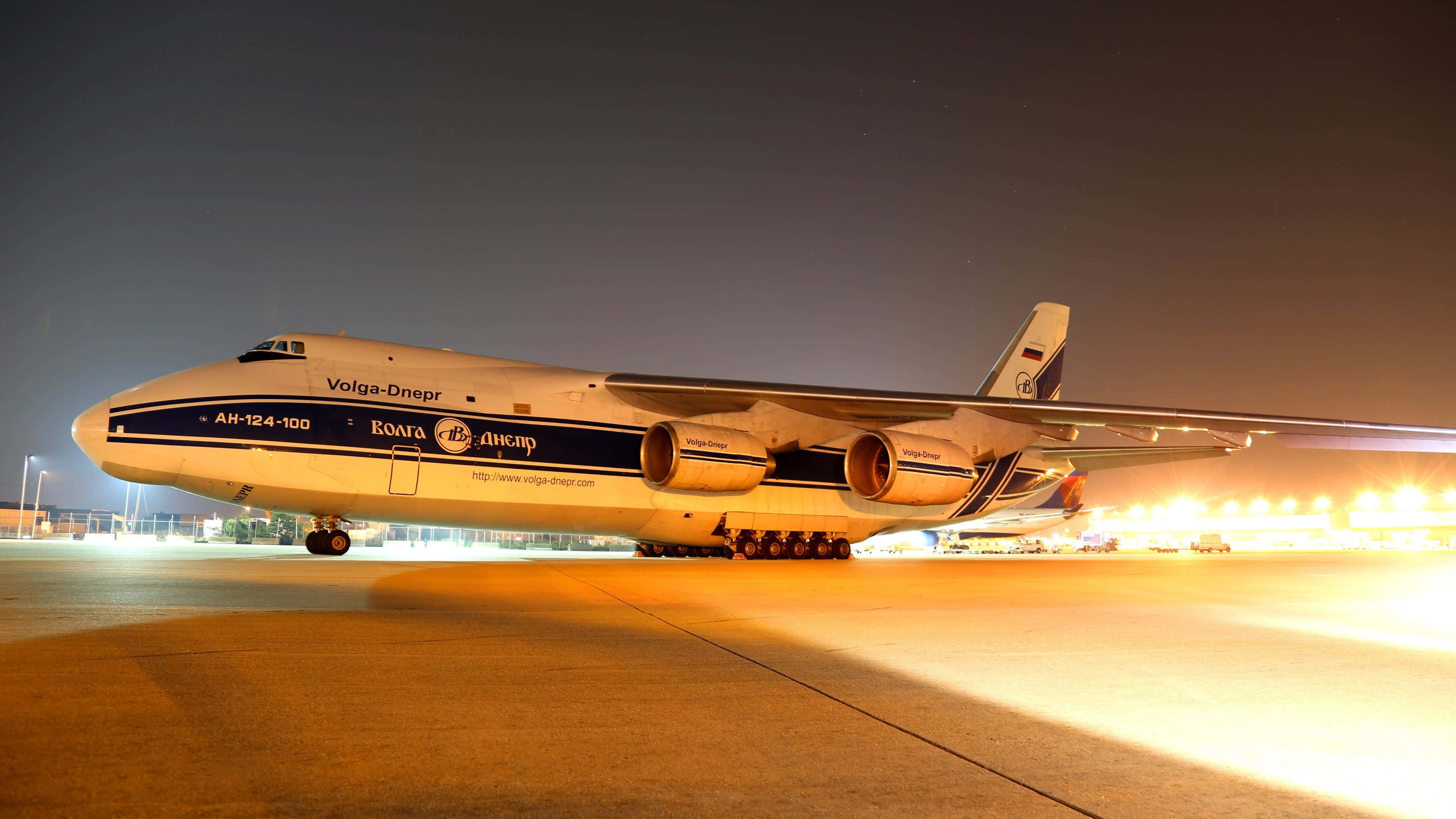 Plane 桌布 壁紙 — 一架巨大的白色 Volga-Dnepr Antonov An-124 貨機在夜間停在跑道上，在黑暗的天空下被溫暖的橙色燈光照亮。