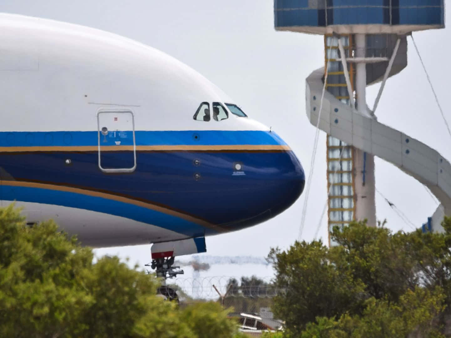 Airbus 桌布 壁紙 — 一架巨大的白色和藍色貨機，機頭圓潤，佔據了畫面主體，部分被綠色樹木和一個高聳的工業塔遮擋，背景是朦朧的天空。