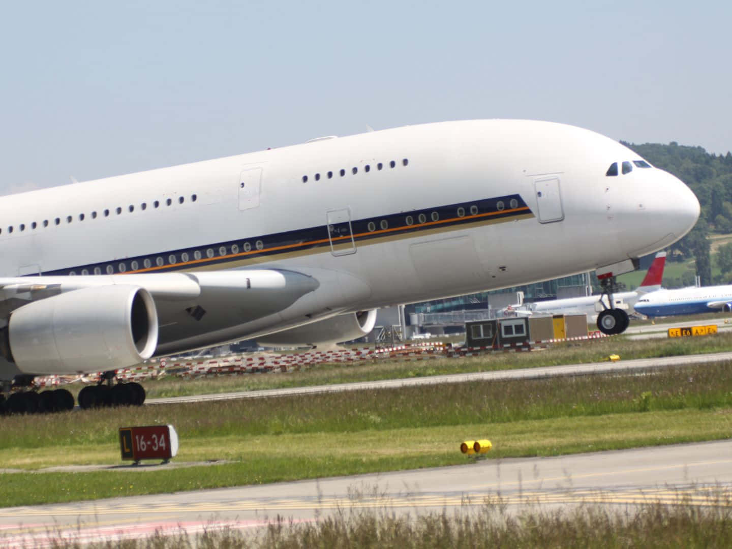 飛機 桌布 壁紙 — 一架巨大的白色空中巴士A380停在機場停機坪上，周圍有綠草和遠處的山丘，天空清澈湛藍。