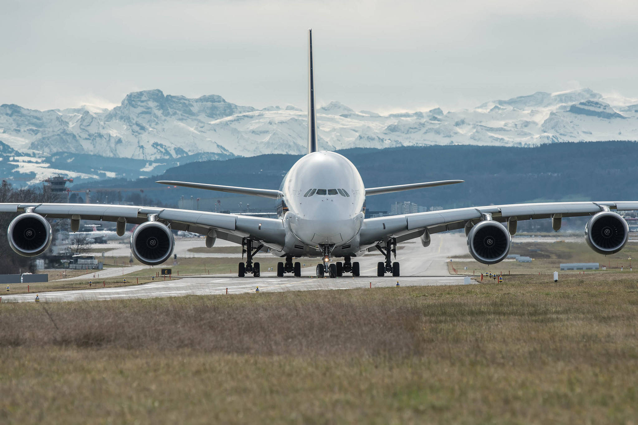 山脈 桌布 壁紙 — 一架巨大的白色空中巴士A380飛機停在跑道上，陰雲密佈的背景中聳立著積雪覆蓋的山脈。