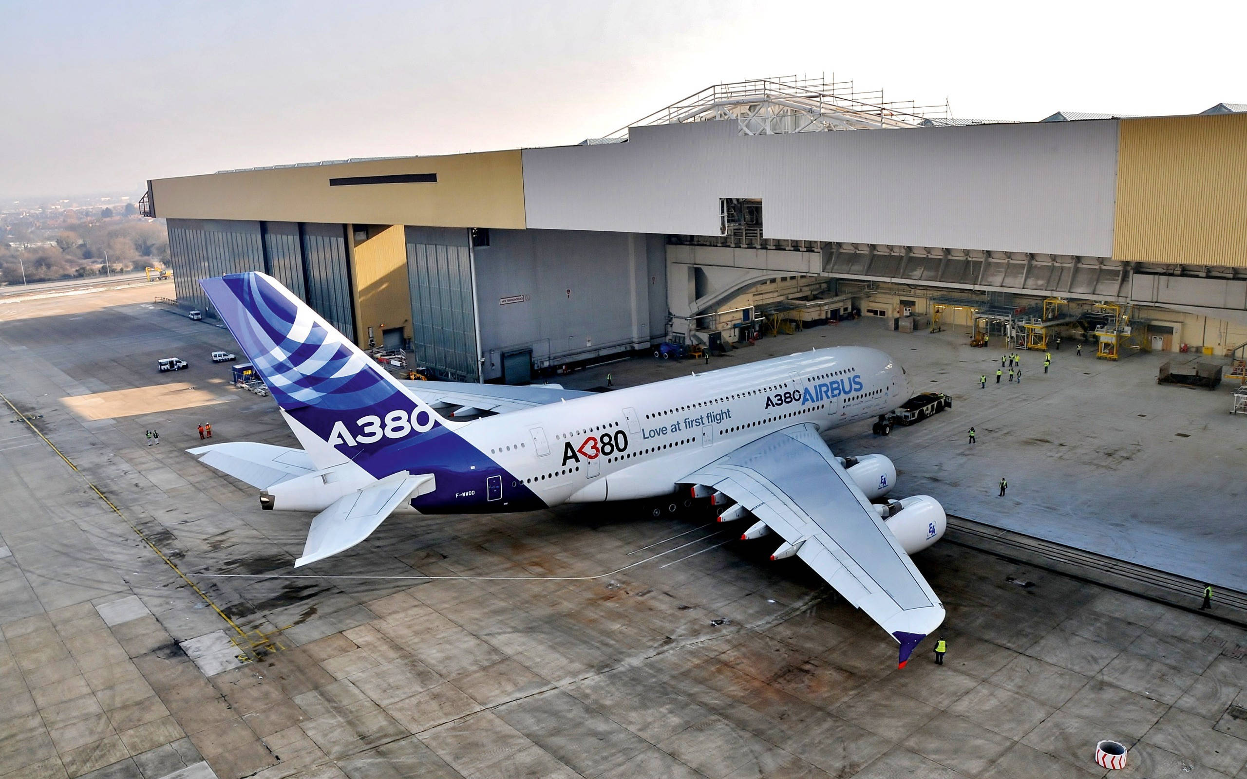 Airport 桌布 壁紙 — 一架巨大的空中巴士A380，帶有「Love at first flight」塗裝，停在機庫外的停機坪上，地勤人員在附近工作。