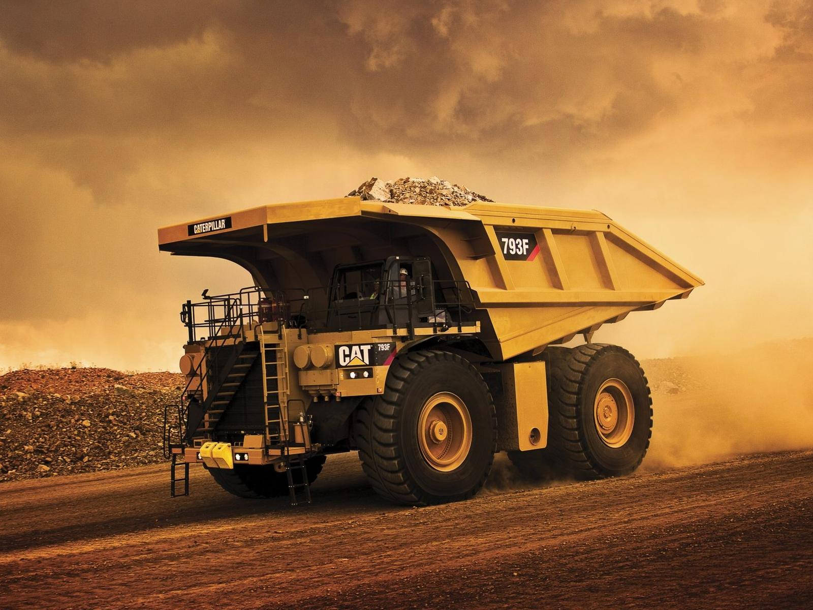 construction 桌布 壁紙 — 一架巨大的黃色Caterpillar 793F礦用自卸車在金色夕陽天空下，穿越塵土飛揚的採石場運送礫石。