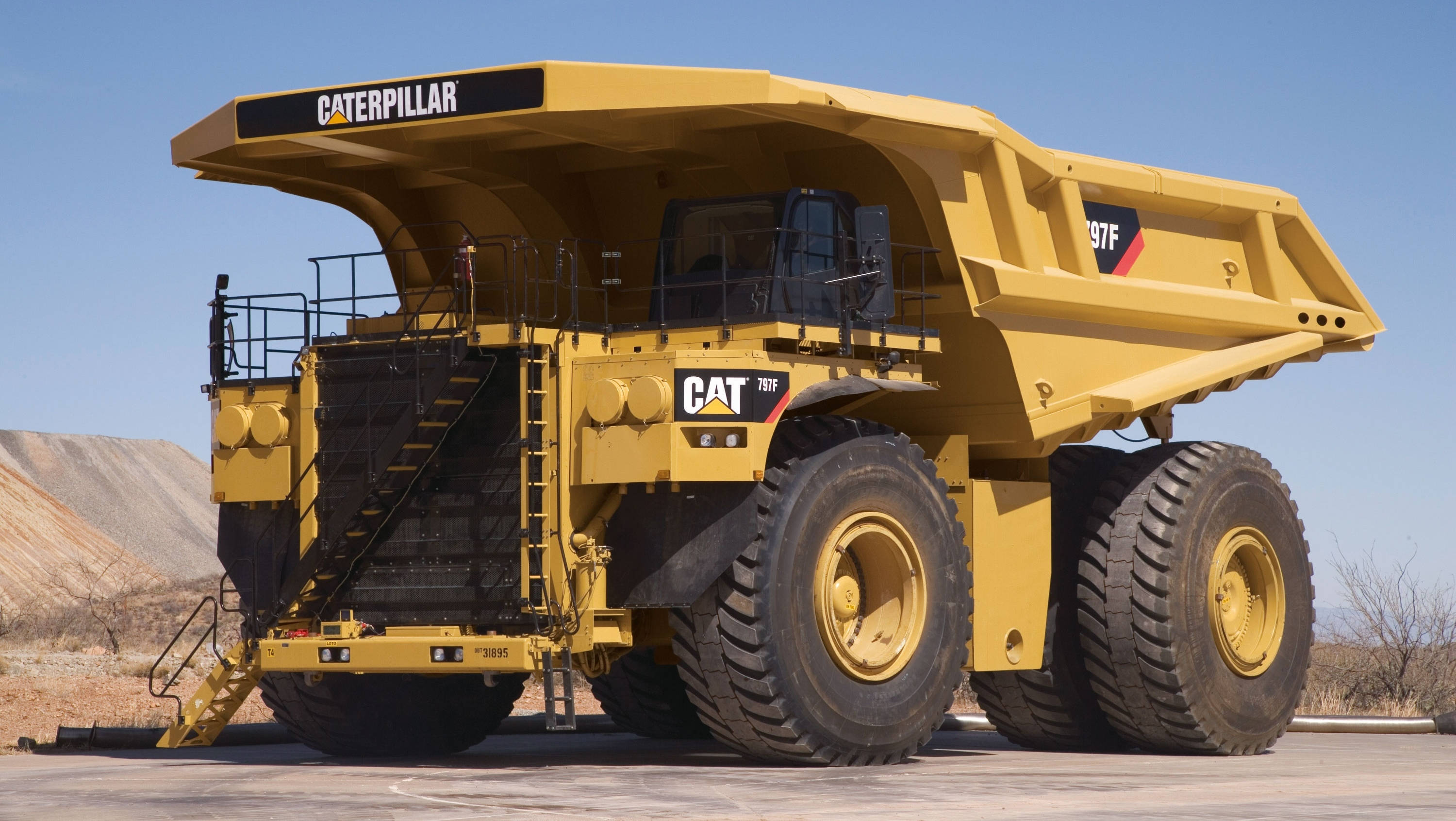Cool Truck 桌布 壁紙 — 一架巨大的黃色Caterpillar 797F礦用自卸車停放在鋪砌路面上，背景是乾旱的山丘和晴朗的藍天。