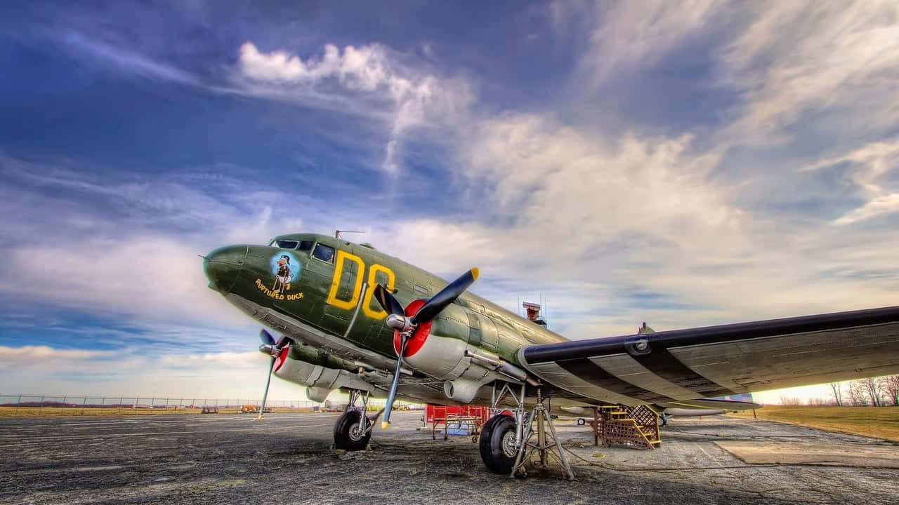 飛機 桌布 壁紙 — 一架帶有 'DC' 標記的復古軍用飛機停在碎石停機坪上，背景是帶有縷縷白雲的戲劇性藍天。