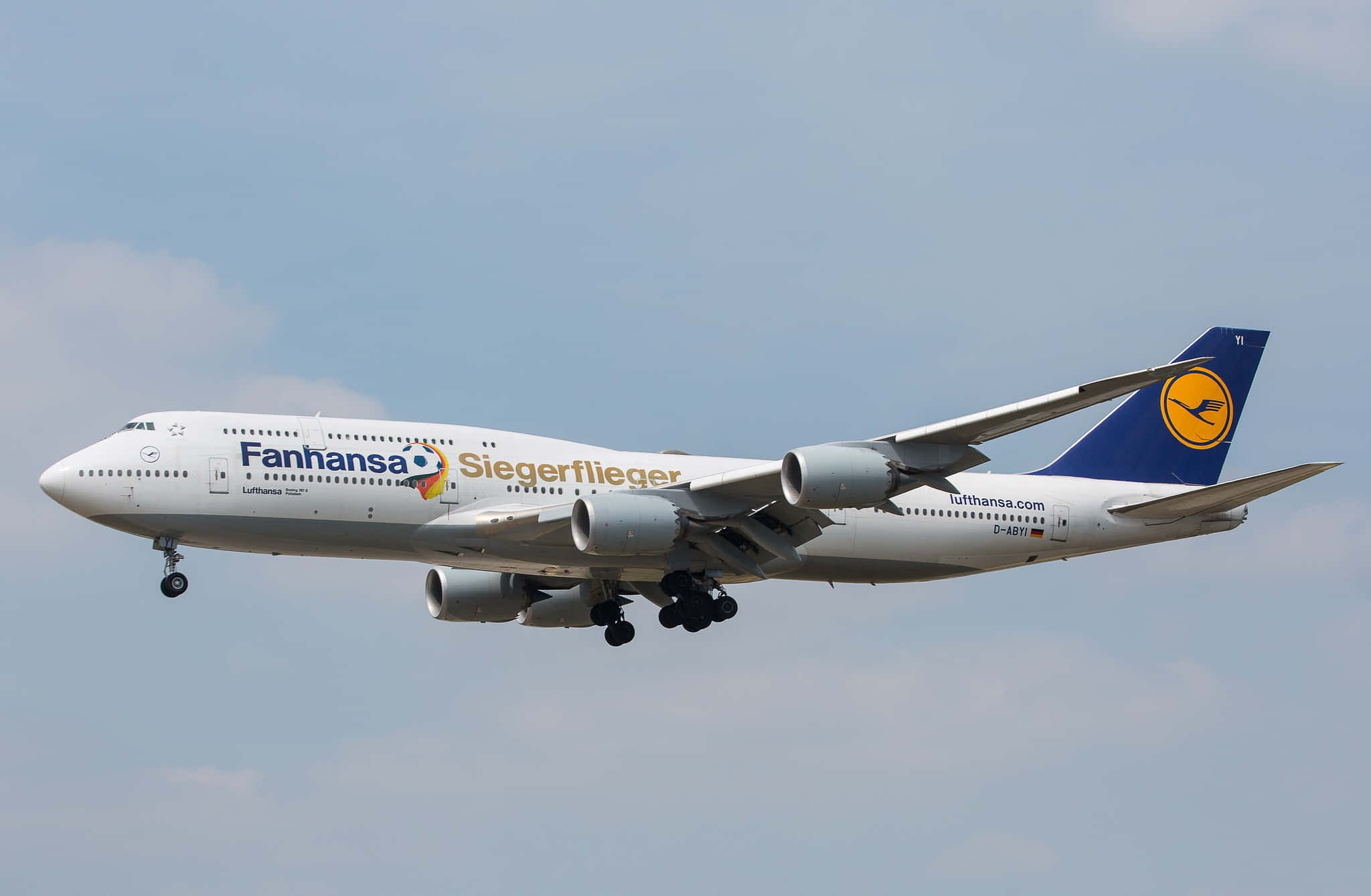 桌布 壁紙 — 一架帶有「Fanhanasa Siegerflieger」塗裝的漢莎航空波音747，在淡藍色天空和零星雲朵中優雅地傾斜飛行。