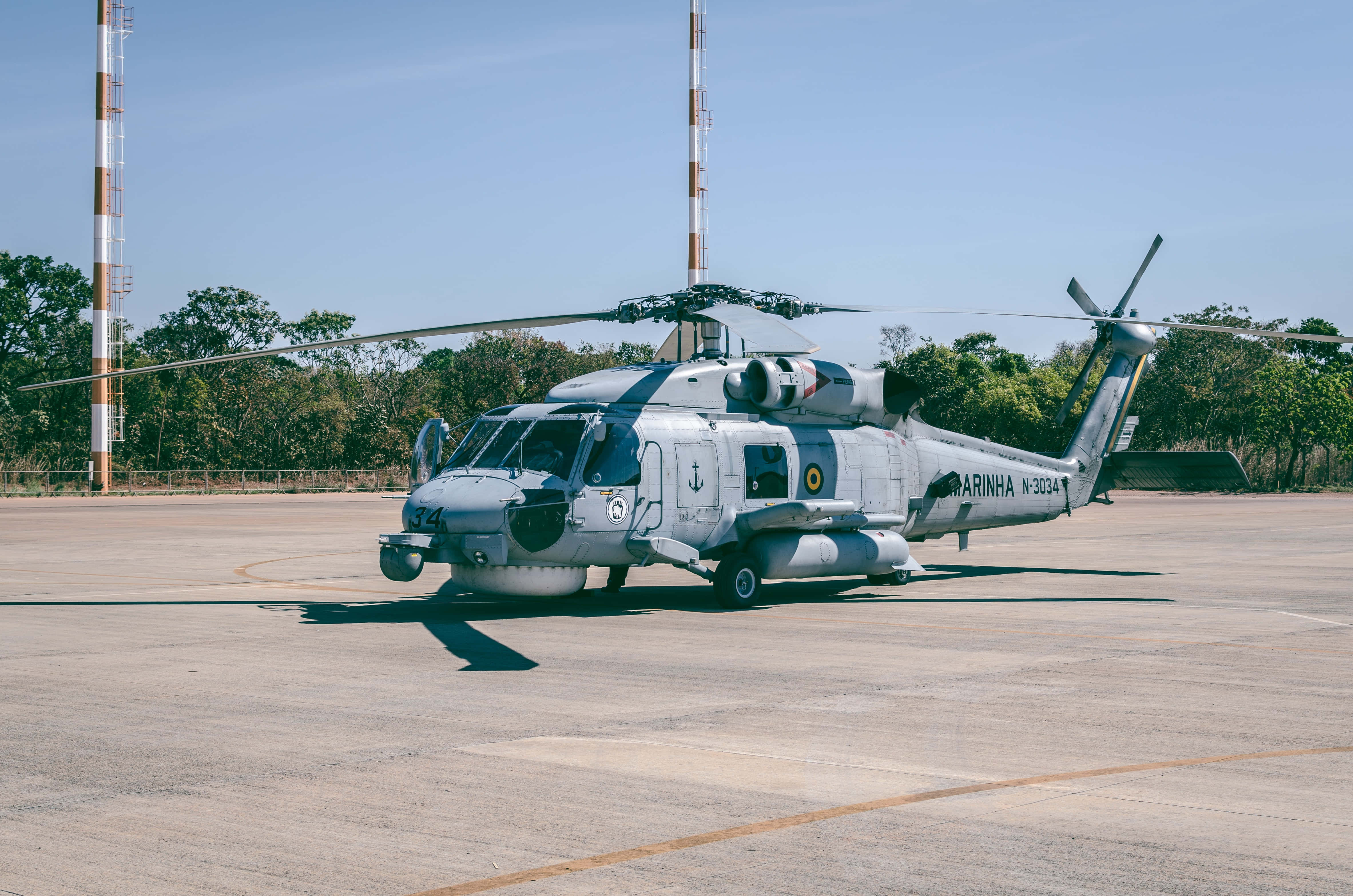 Helicopter 桌布 壁紙 — 一架帶有 'MARINHA' 標誌的灰色軍用直升機停在停機坪上，背景是晴朗的藍天、樹木和通訊塔。
