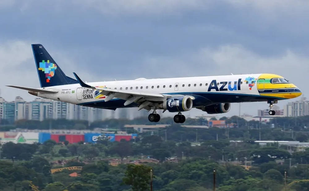 飛機 桌布 壁紙 — 一架帶有像素化洗拿（Ayrton Senna）塗裝的藍色航空（Azul Airlines）飛機，在陰雲密佈的天空下，降落在綠色城市景觀之上。