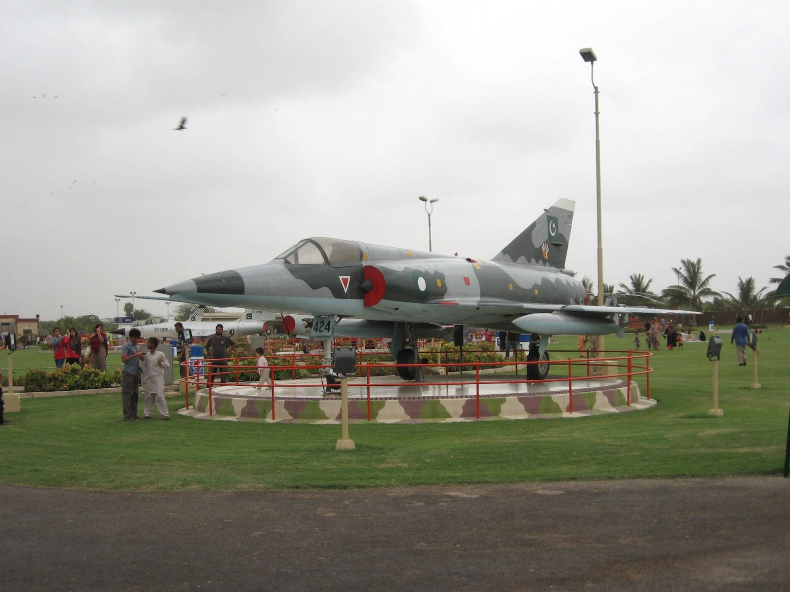 Karachi 桌布 壁紙 — 一架帶有巴基斯坦標誌的灰色軍用噴射機，在陰天之下，立於草地戶外博物館的圓形基座上，周圍有參觀者。