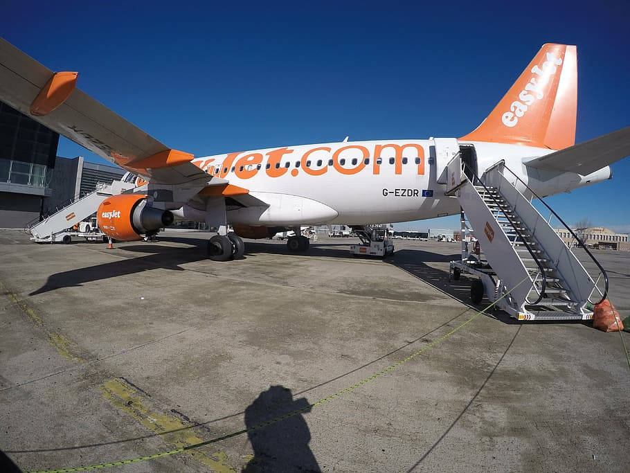 Easyjet 桌布 壁紙 — 一架帶有橙色點綴的易捷航空飛機停在機場閘口，附有登機梯，背景是晴朗的藍天。