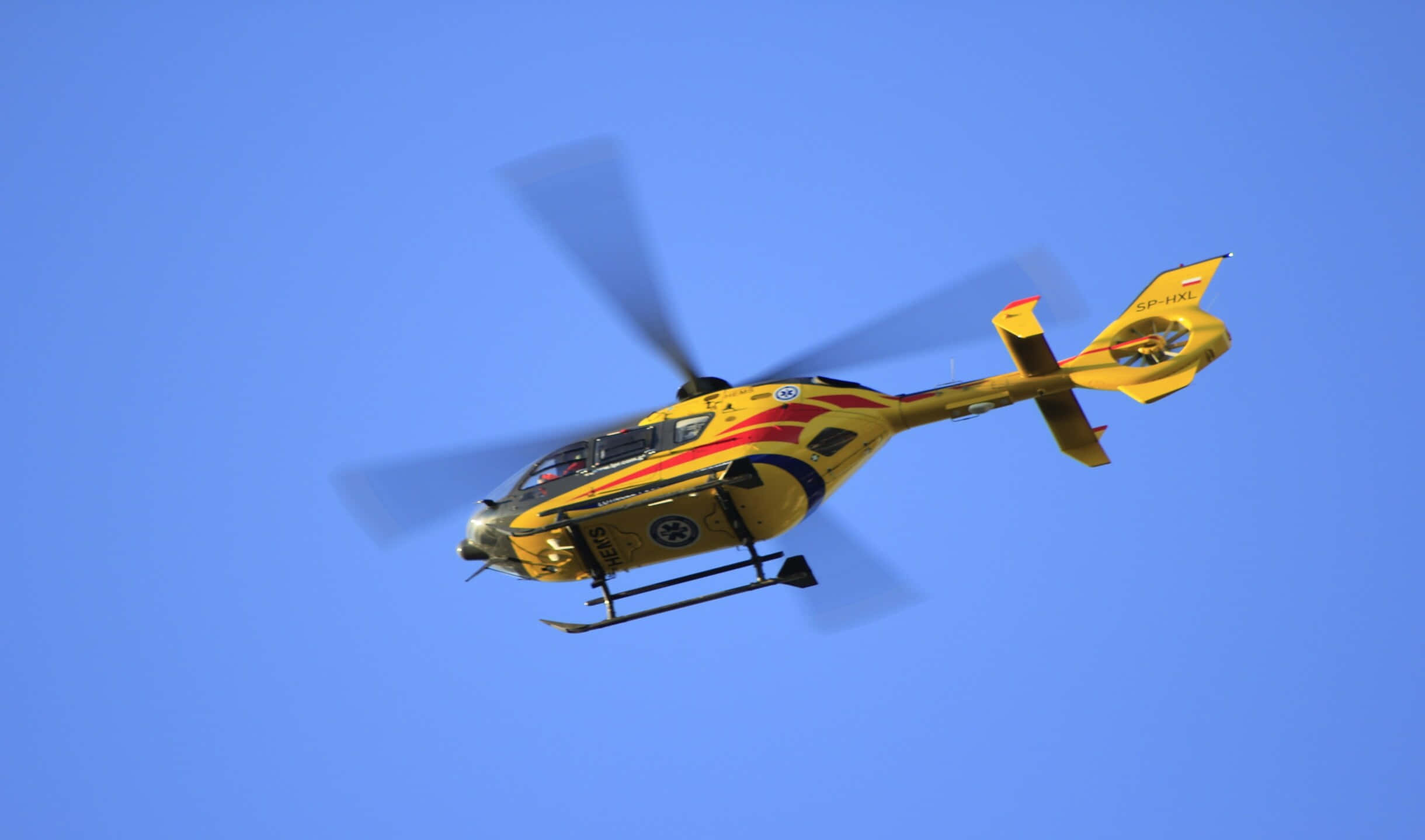 2440x1440 Helicopters 桌布 壁紙 — 一架帶有紅色火焰貼紙的亮黃色救援直升機在萬里無雲的藍天中翱翔。