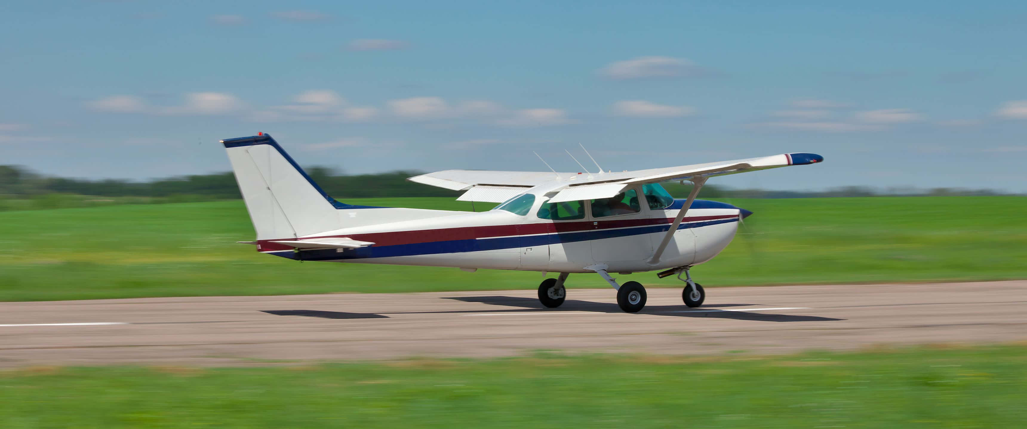 3440x1440p Small Planes 桌布 壁紙 — 一架帶有紅藍條紋的白色小型螺旋槳飛機在跑道上滑行，背景是晴朗的藍天和零星的白雲。