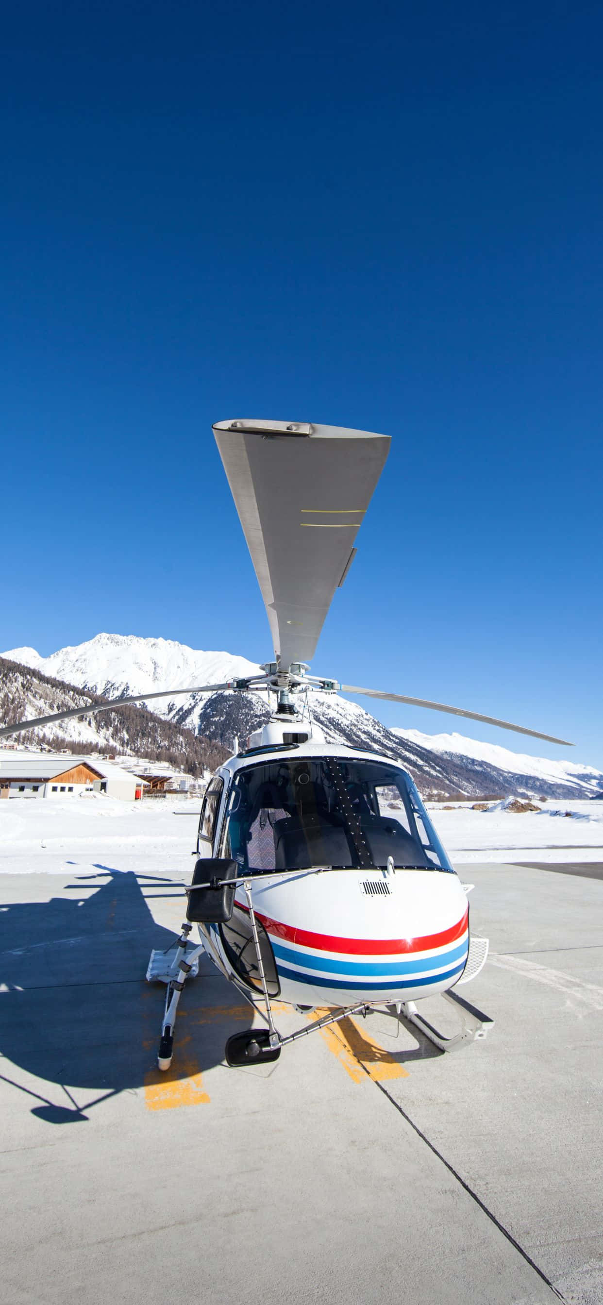 iPhone Xs Max Helicopters 桌布 壁紙 — 一架帶有紅藍條紋的白色直升機停在積雪的停機坪上，背景是白雪覆蓋的山脈和清澈的藍天。
