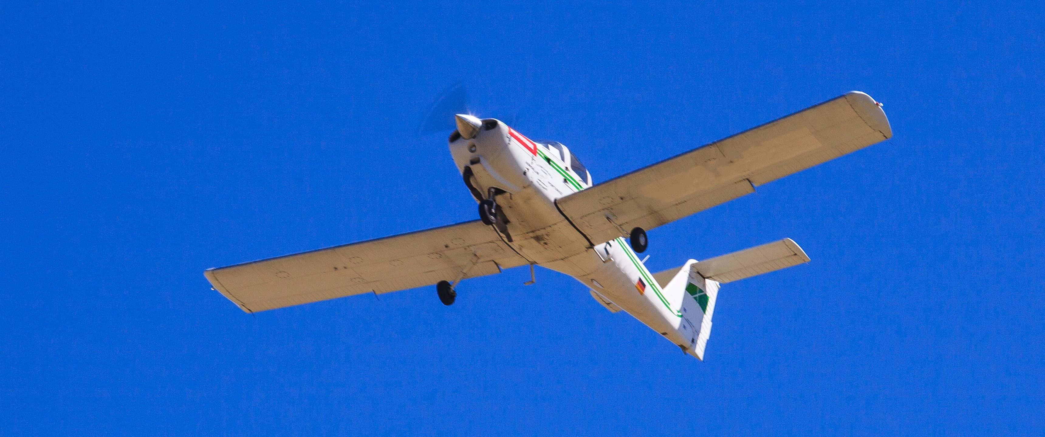 3440x1440p Small Planes 桌布 壁紙 — 一架帶有綠色和紅色條紋的白色小型單引擎飛機在鮮豔的藍天中翱翔。
