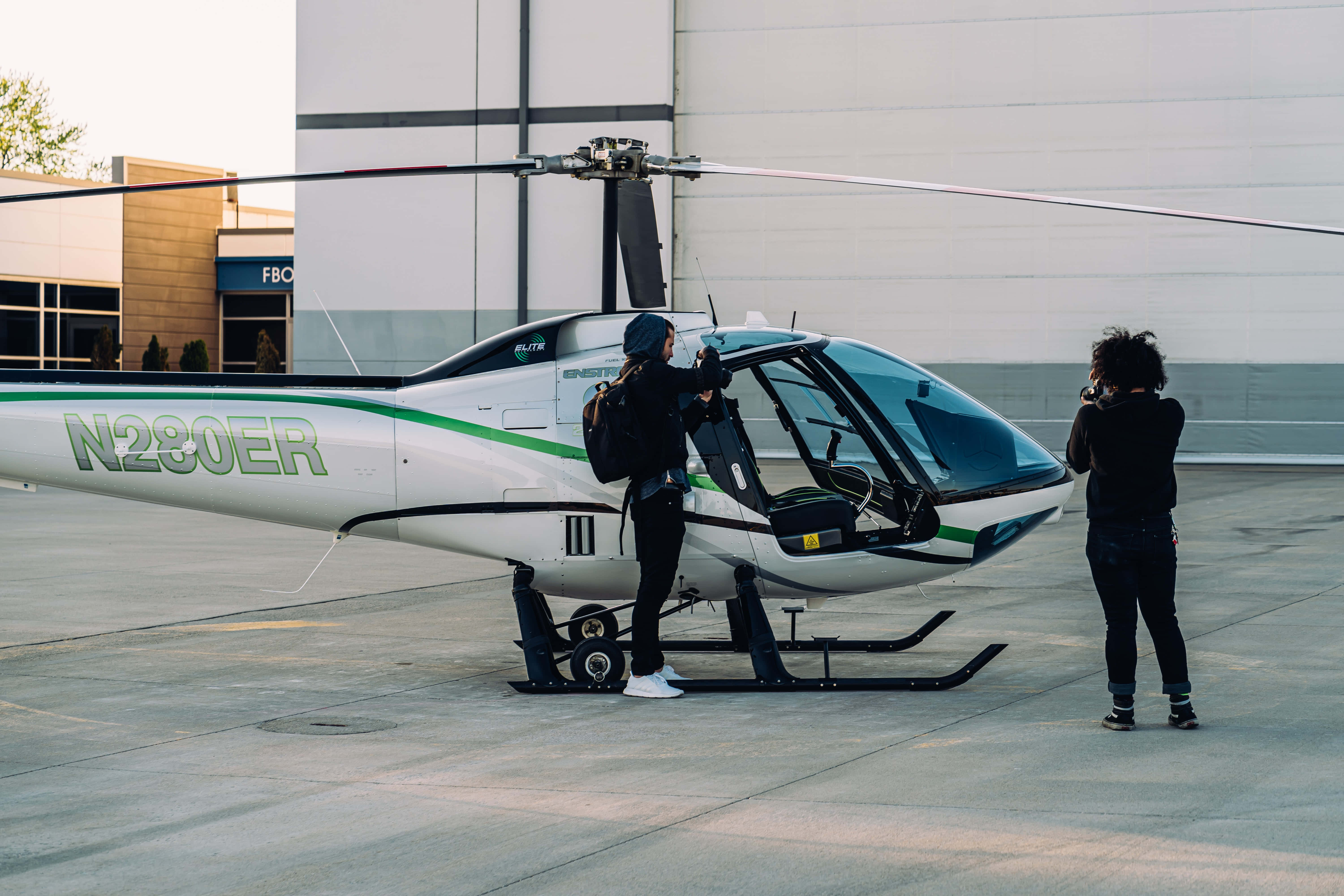 Helicopter 桌布 壁紙 — 一架帶有綠色點綴的時尚白色直升機停在停機坪上，兩人在機庫附近準備飛行。