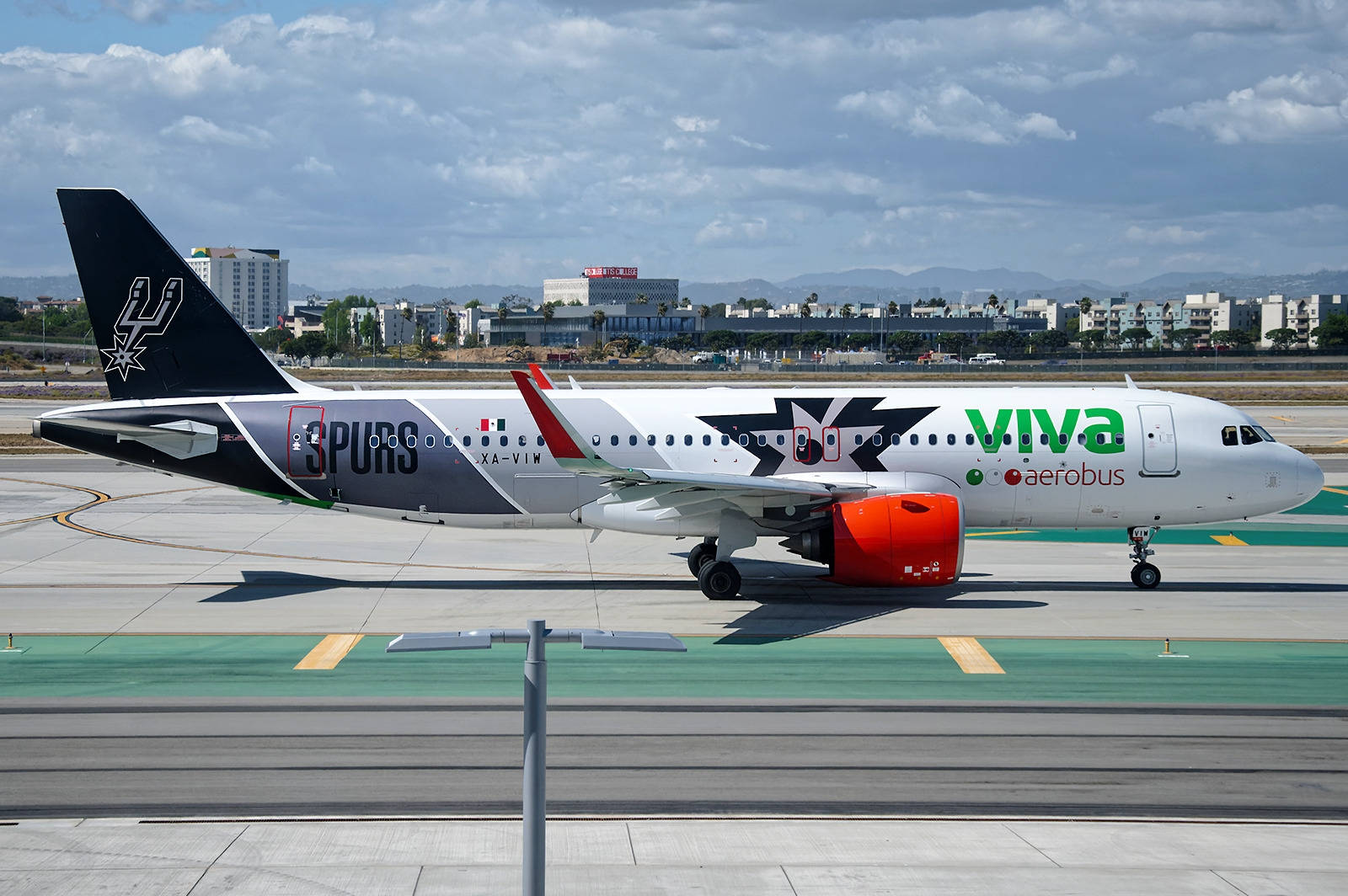 飛機 桌布 壁紙 — 一架帶有聖安東尼奧馬刺隊塗裝的Viva Aerobus空中巴士A320在部分多雲的天空下，於陽光明媚的機場停機坪上滑行。