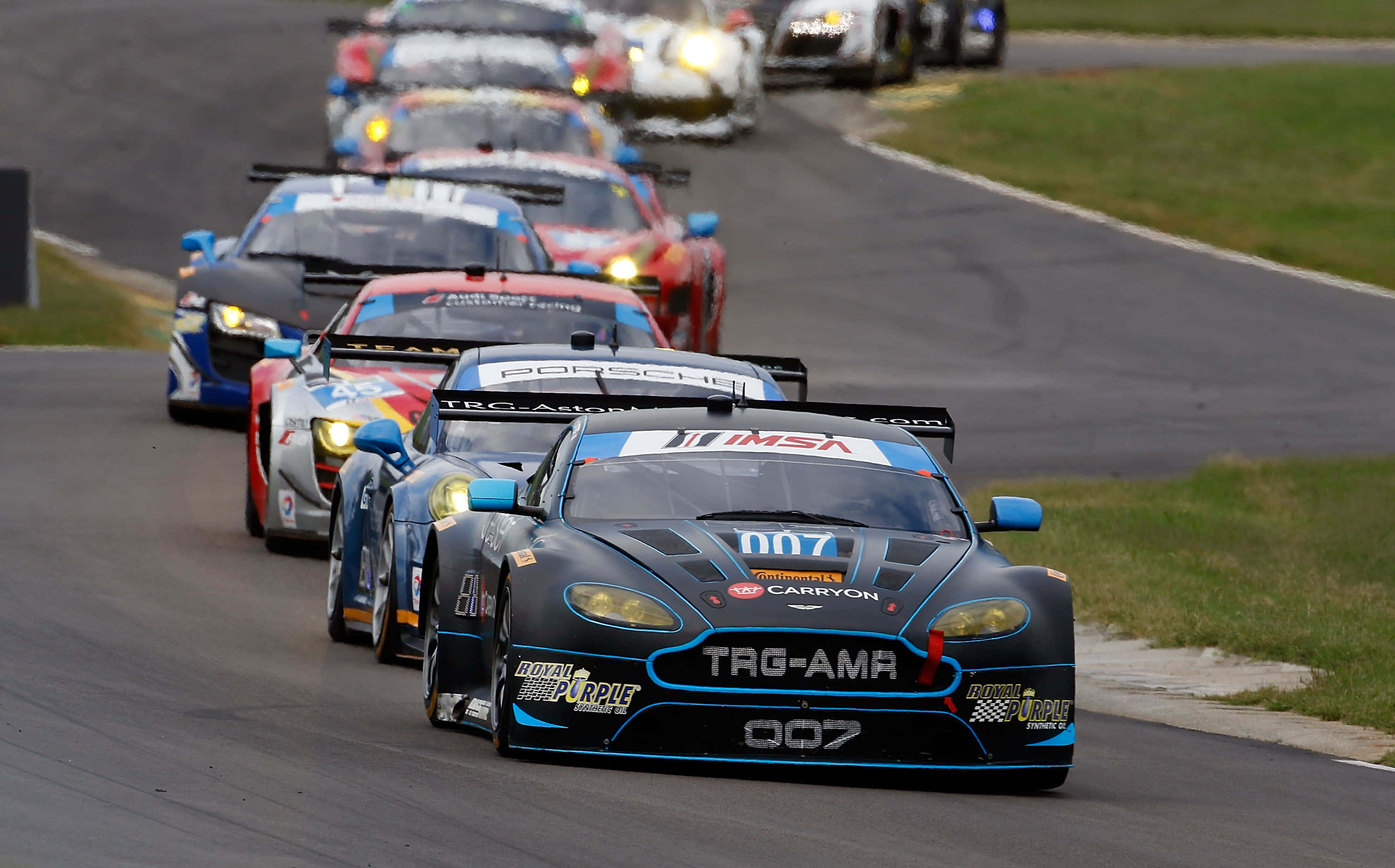 Car Racing 桌布 壁紙 — 一架帶有藍色點綴的時尚黑色 Aston Martin 賽車，在黃昏時分帶領一群色彩繽紛的跑車在蜿蜒的瀝青賽道上馳騁。