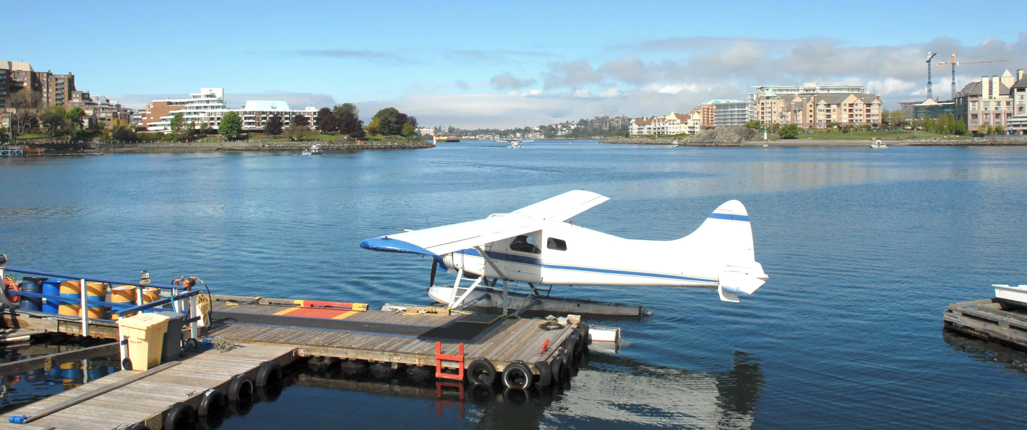3440x1440p Small Planes 桌布 壁紙 — 一架帶有藍色點綴的白色水上飛機停泊在木製碼頭上，平靜的藍色水面上，遠處岸邊排列著城市建築，天空部分多雲。