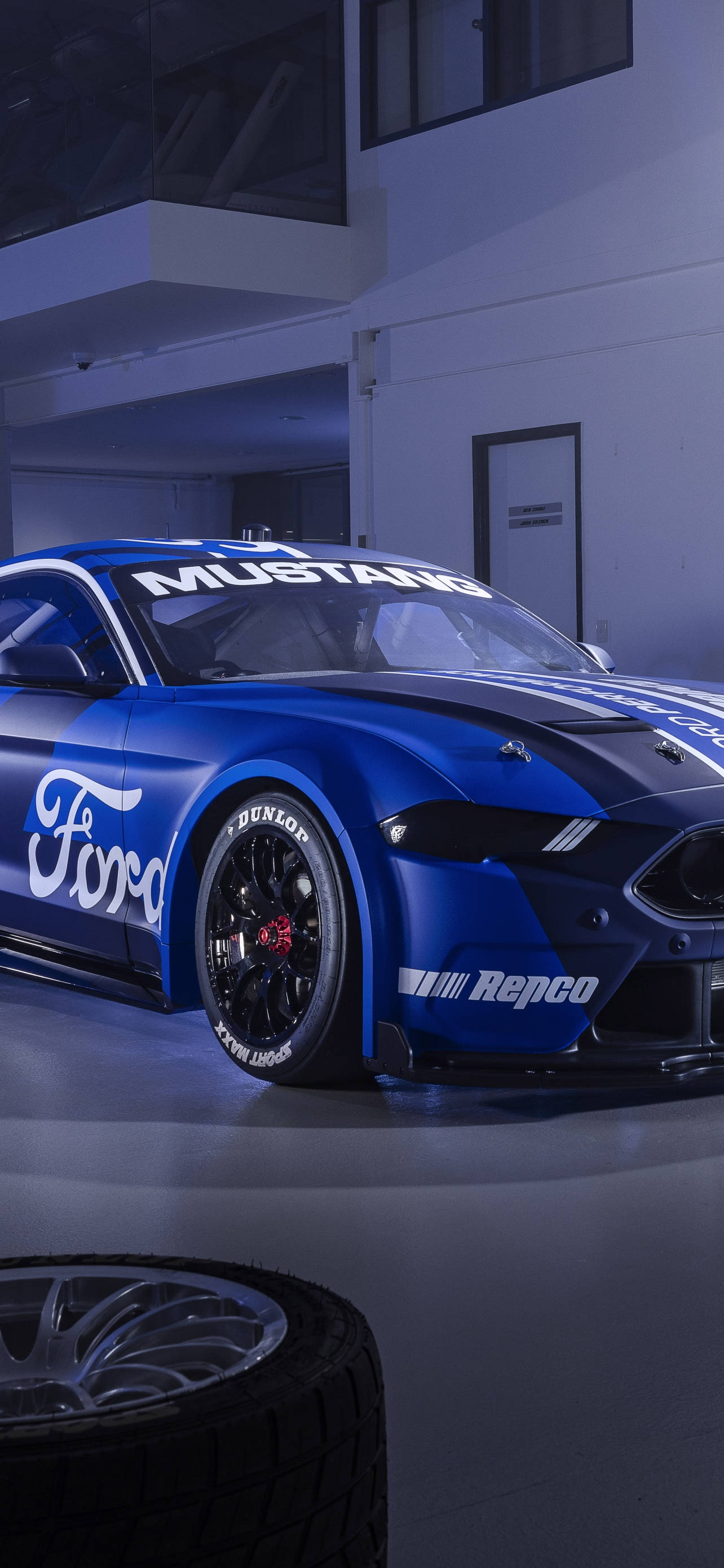 Cool Mustang 桌布 壁紙 — 一架帶有贊助商貼紙的亮藍色福特野馬賽車，在冷藍色燈光下停放在室內，前景有一個備用輪胎。