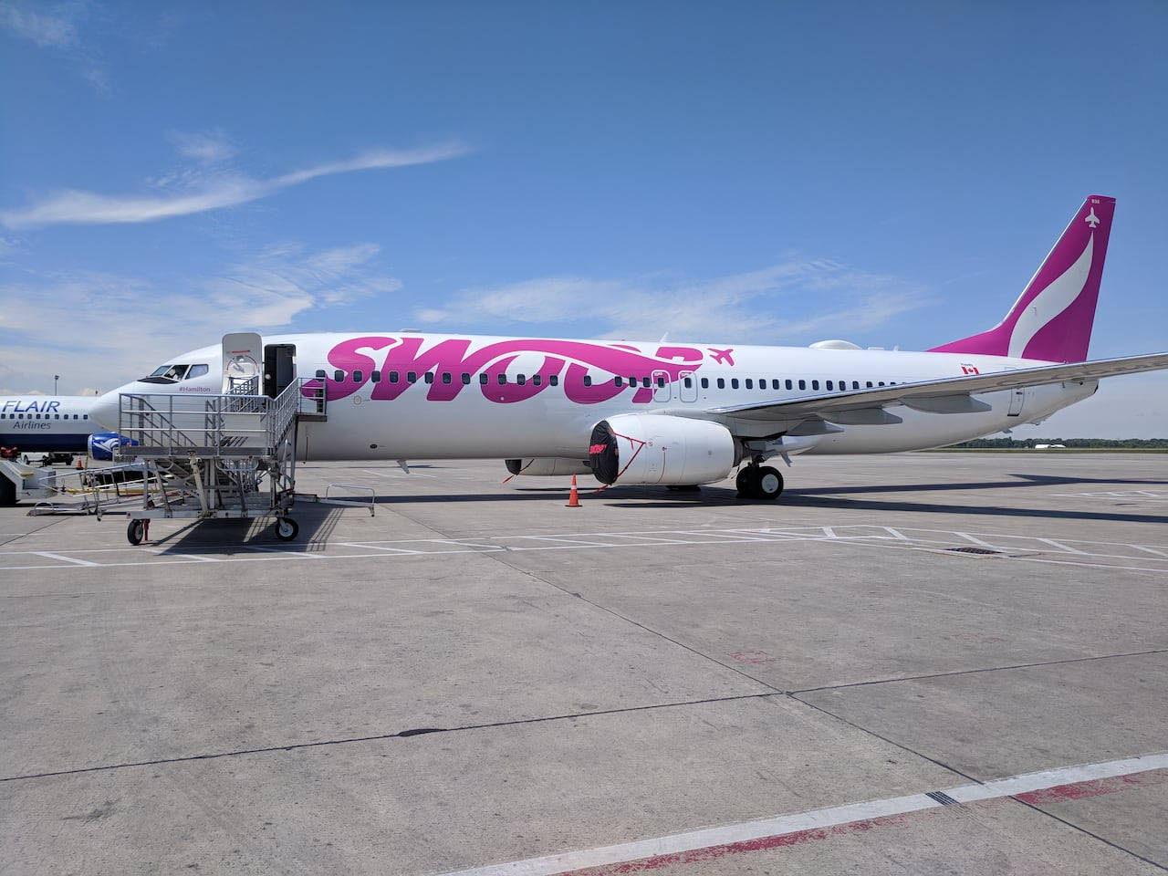 Airport 桌布 壁紙 — 一架帶有醒目粉紅色品牌的白色Swoop Airlines噴射機停在陽光明媚的停機坪上，藍天白雲，並連接了乘客登機橋。