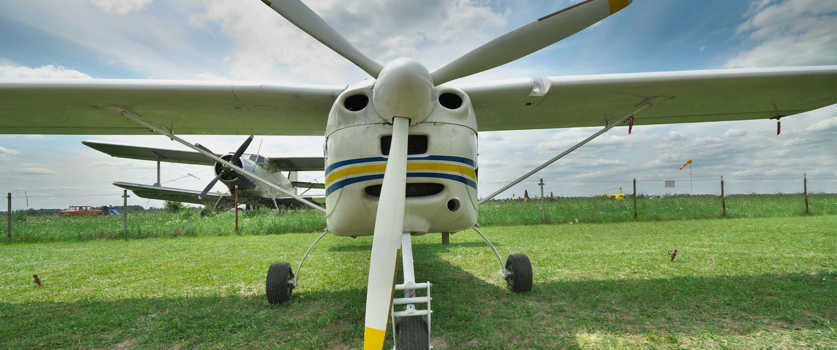 3440x1440p Small Planes 桌布 壁紙 — 一架帶有黃色和藍色條紋的復古單引擎飛機停在草地上，天空部分多雲。