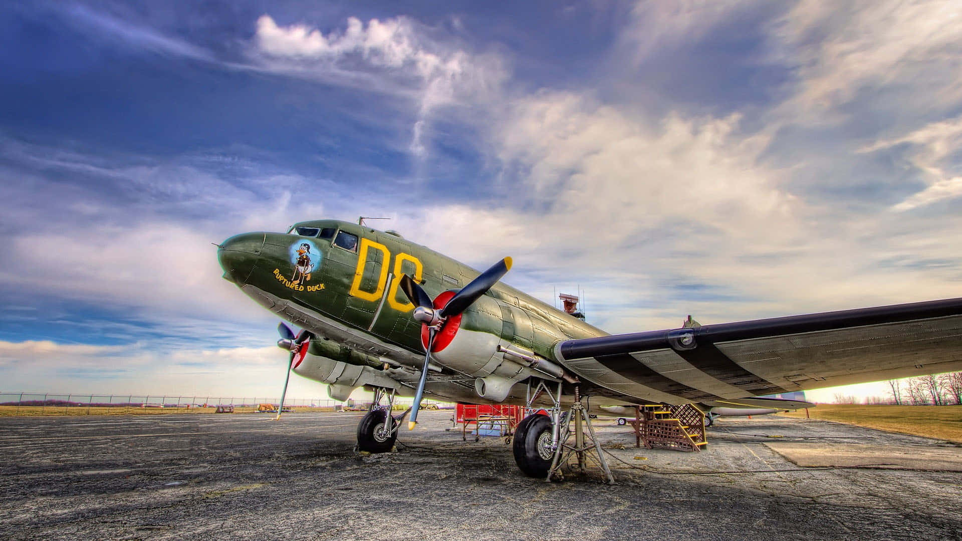 HD Plane 桌布 壁紙 — 一架帶有黃色標記的復古綠色軍用飛機停在停機坪上，背景是帶有縷縷白雲的戲劇性藍天。
