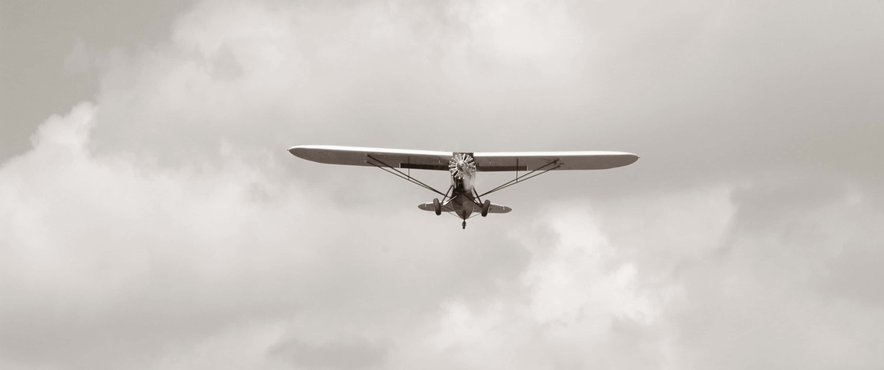 3440x1440p Small Planes 桌布 壁紙 — 一架復古單翼機在濃密多雲的天空中翱翔，光線柔和擴散。