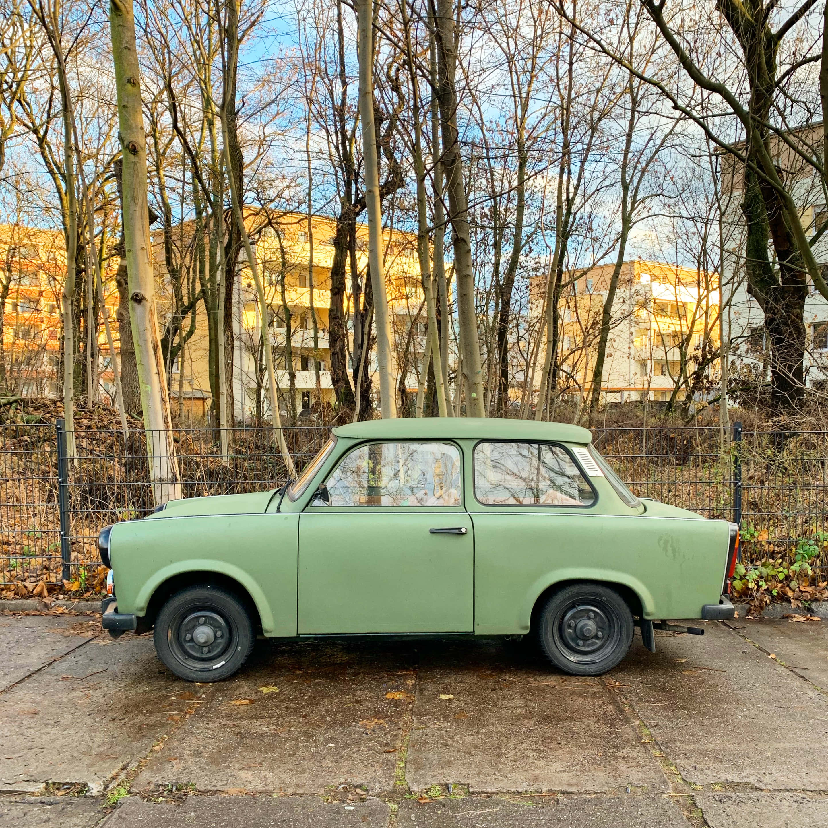 Vintage Car 桌布 壁紙 — 一架復古橄欖綠色的Trabant汽車停放在混凝土場地上，周圍是光禿禿的樹木和住宅建築，天空清澈湛藍。