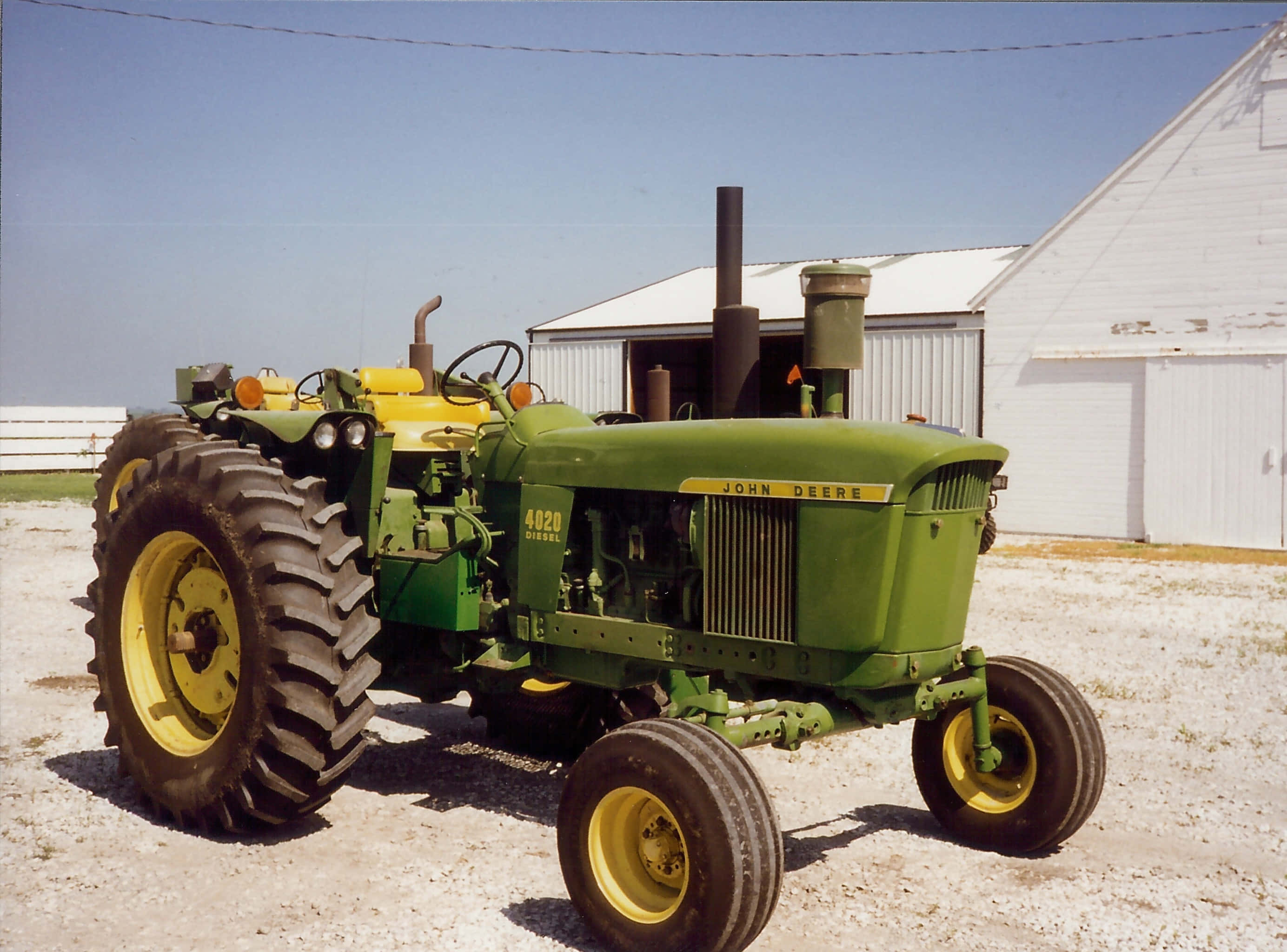 Tractor 桌布 壁紙 — 一架復古綠色和黃色的John Deere 4020柴油拖拉機停泊在白色穀倉前的碎石地上，背景是晴朗的藍天。