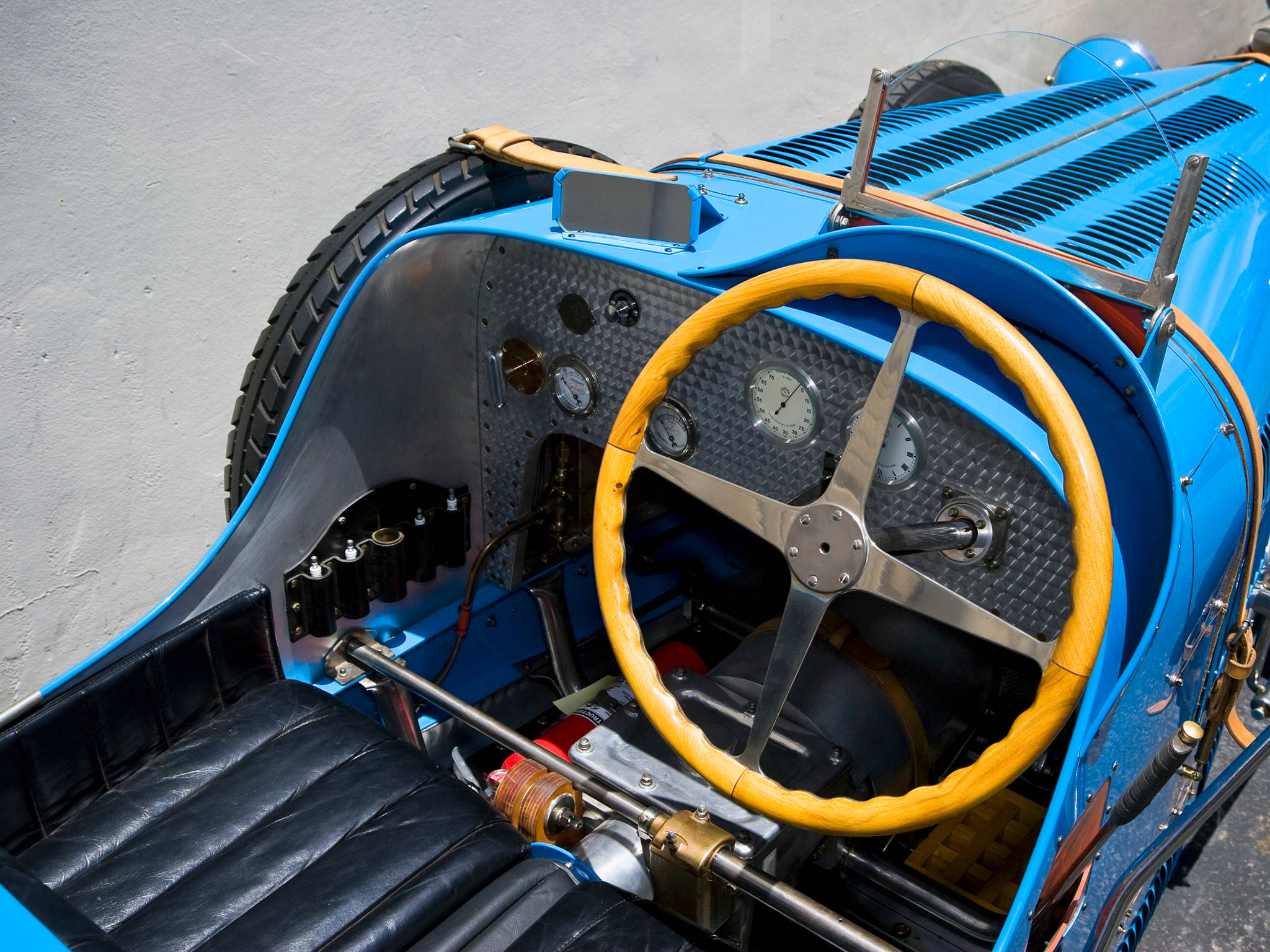 bugatti iPhone 桌布 壁紙 — 一架復古藍色賽車的駕駛艙配備木製方向盤、黑色皮革座椅和暴露的機械儀表，背景為素牆。