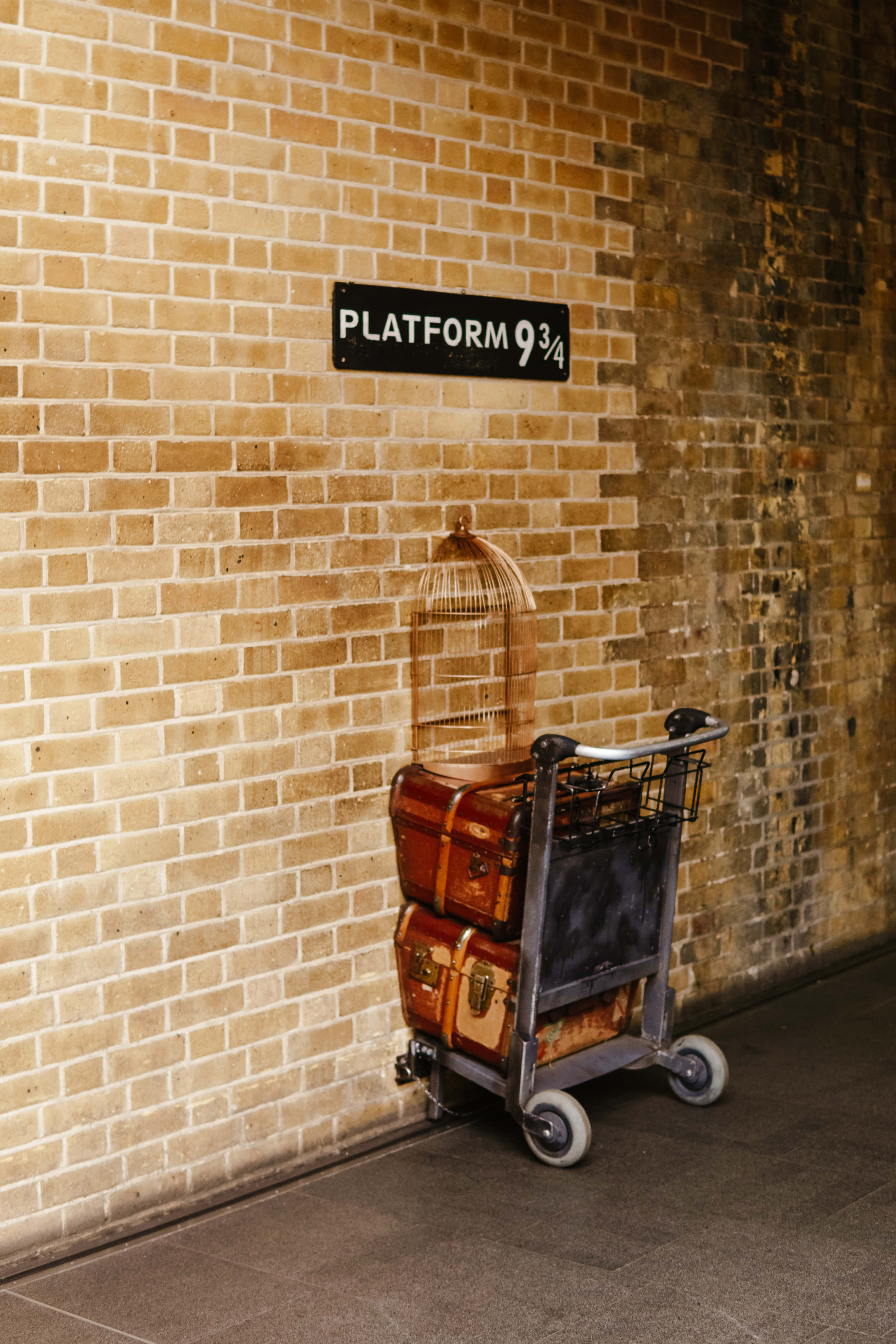Platform 9 3/4 桌布 壁紙 — 一架復古行李車堆滿舊皮箱和鳥籠，立在磚牆旁，上方有「Platform 9¾」標誌。