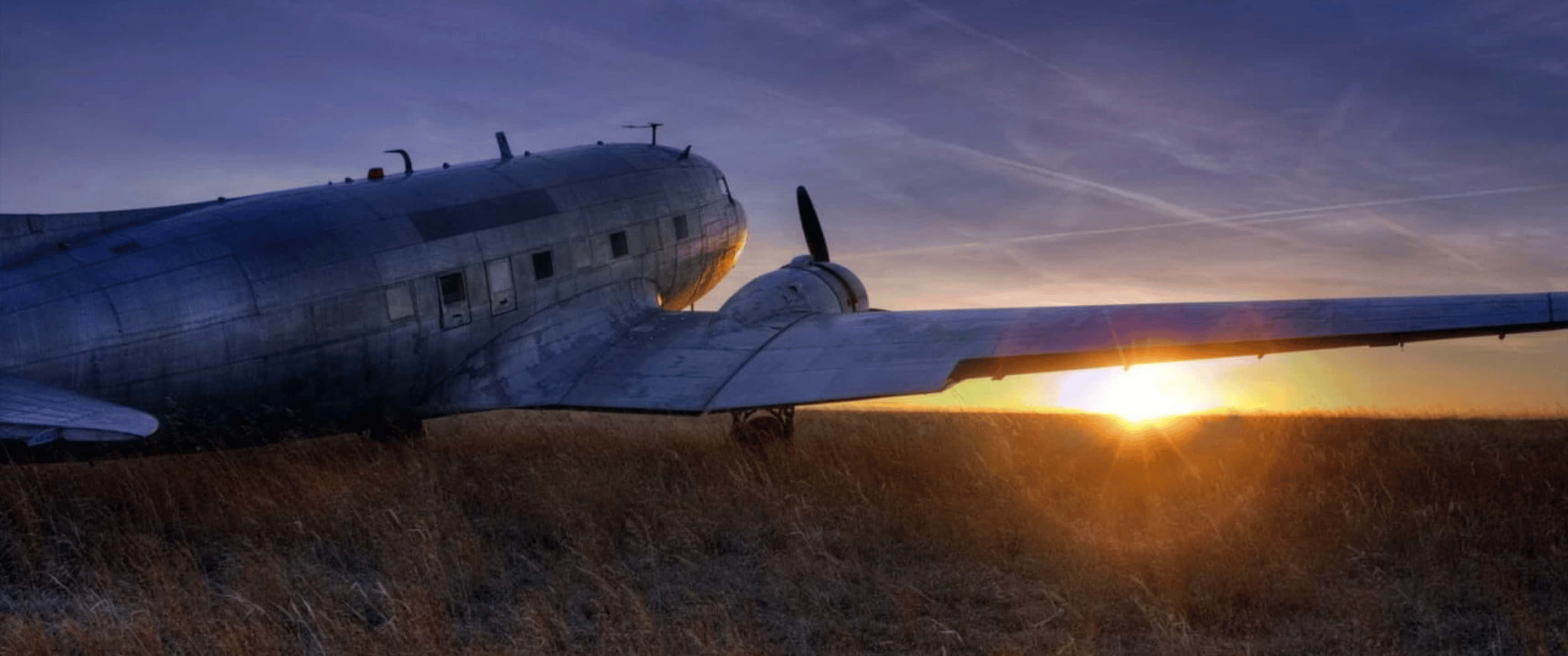 Plane 桌布 壁紙 — 一架復古銀色飛機停放在乾草田野中，夕陽在它身後落下，散發出溫暖的金色光芒。