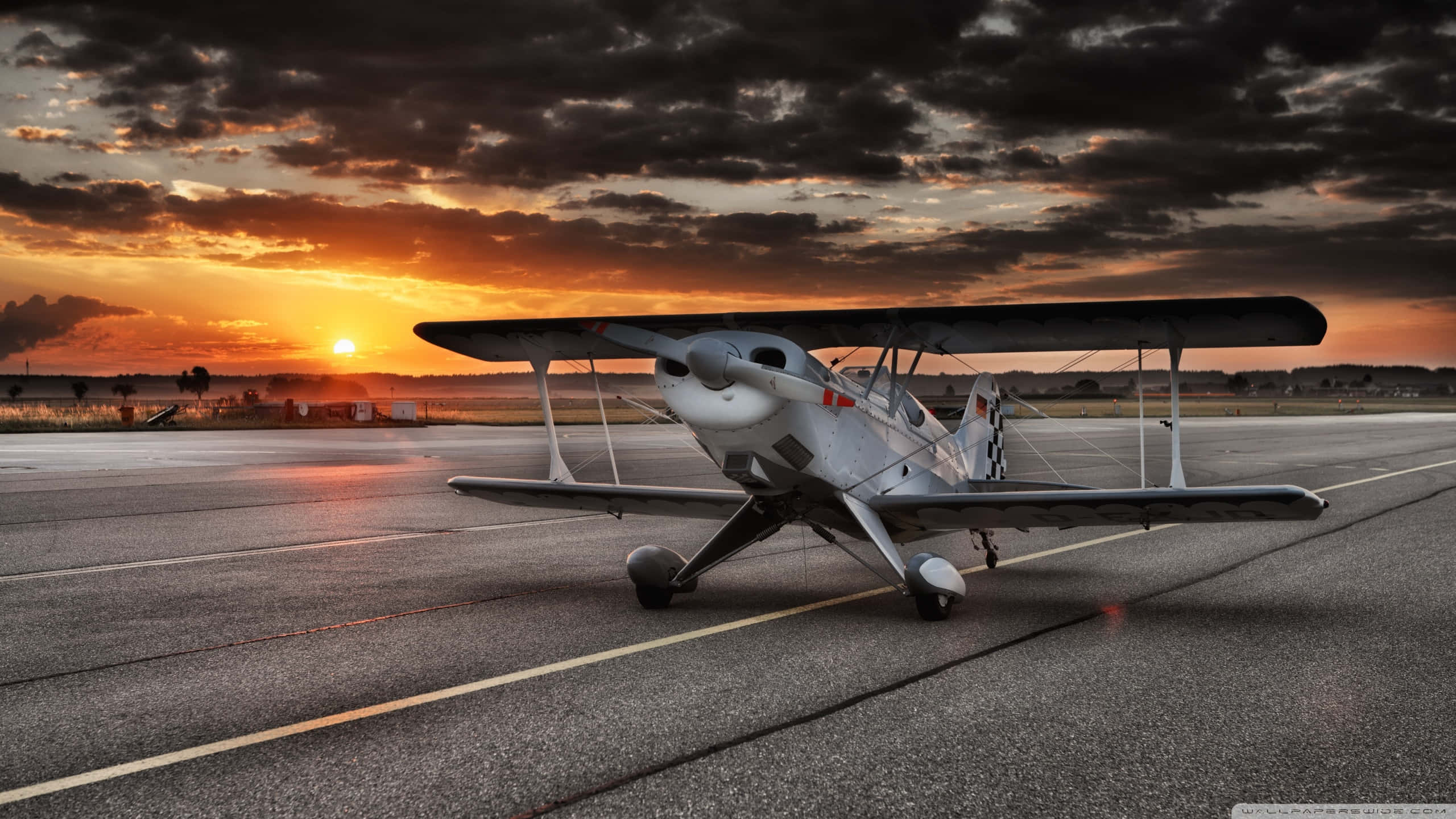 1440p Small Planes 桌布 壁紙 — 一架復古雙翼飛機停在機場停機坪上，熾熱的夕陽用橙色和烏雲塗滿天空。