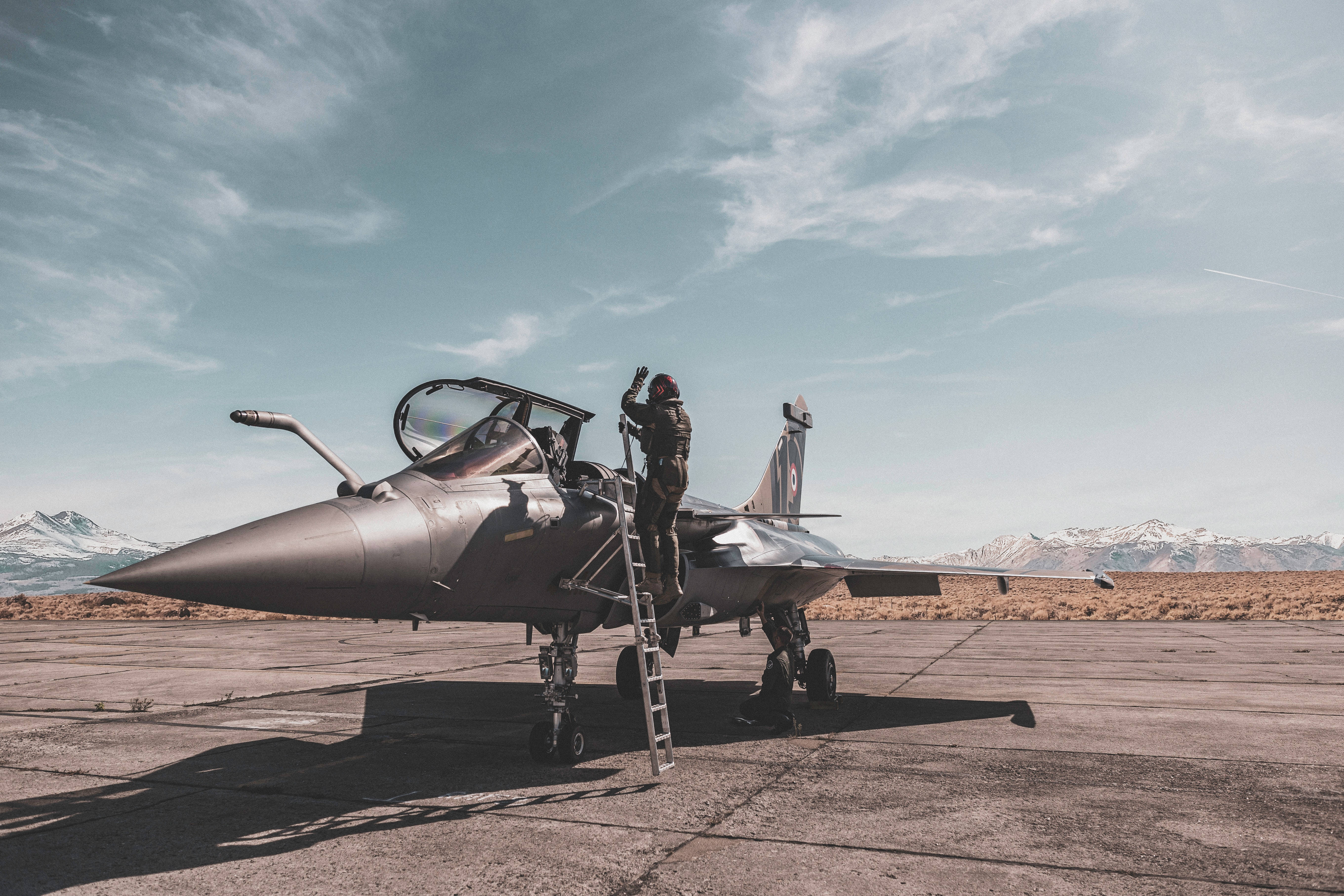 軍事動圖 桌布 壁紙 — 一架戰鬥機停在停機坪上，一名飛行員正在攀爬梯子，背景是白雪覆蓋的山脈和部分多雲的天空。