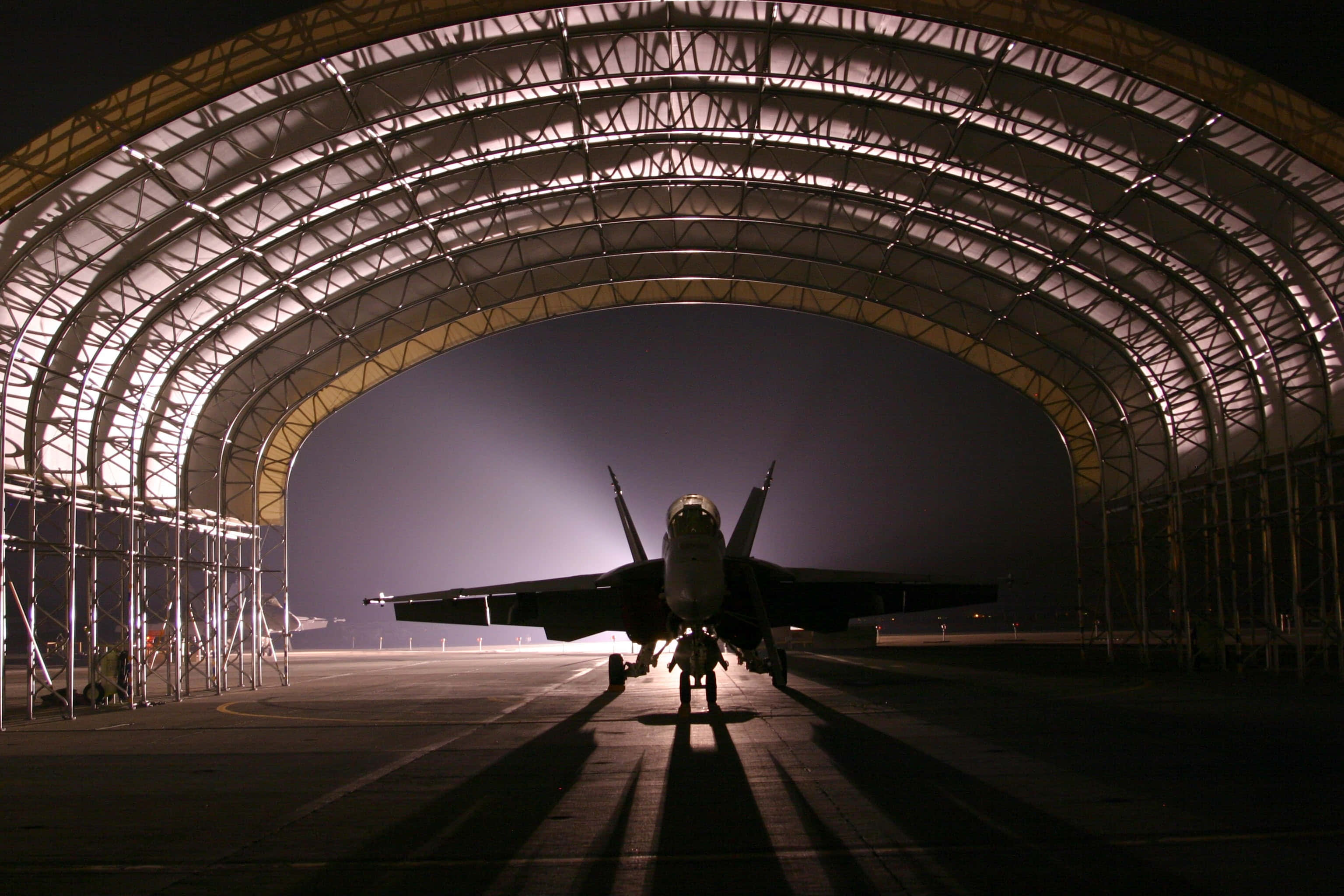 Military Plane 桌布 壁紙 — 一架戰鬥機在夜間於巨大機庫內呈現剪影，戲劇性的燈光投射出長長的陰影。