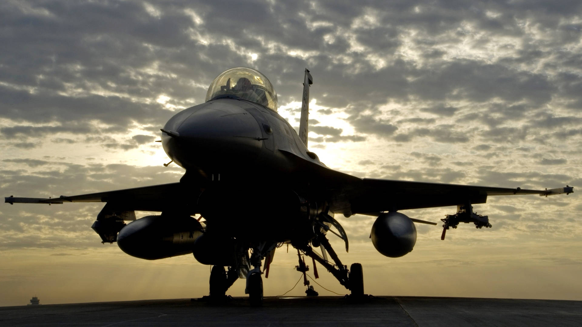 Military Jets 桌布 壁紙 — 一架戰鬥機在戲劇性的多雲日落天空背景下形成剪影，可見駕駛艙和掛載武器。