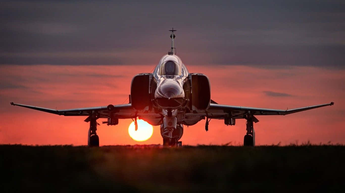 日落 桌布 壁紙 — 一架戰鬥機面向鏡頭，夕陽在其後方落下，在天空中投射出熾熱的橙色光芒。
