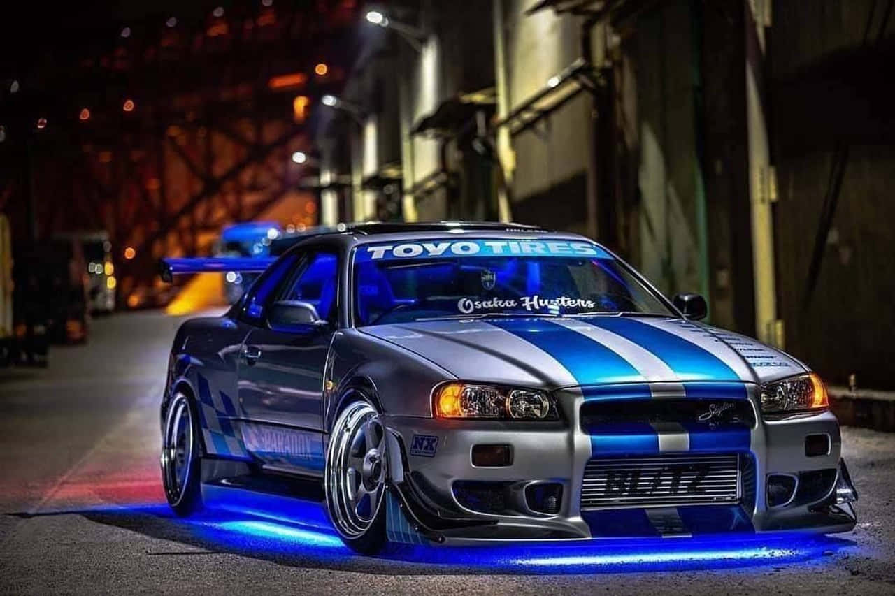 Paul Walker Skyline 桌布 壁紙 — 一架改裝的日產Skyline GTR，帶有藍色和銀色條紋，在夜間於粗糙的城市小巷中霓虹燈下發光。