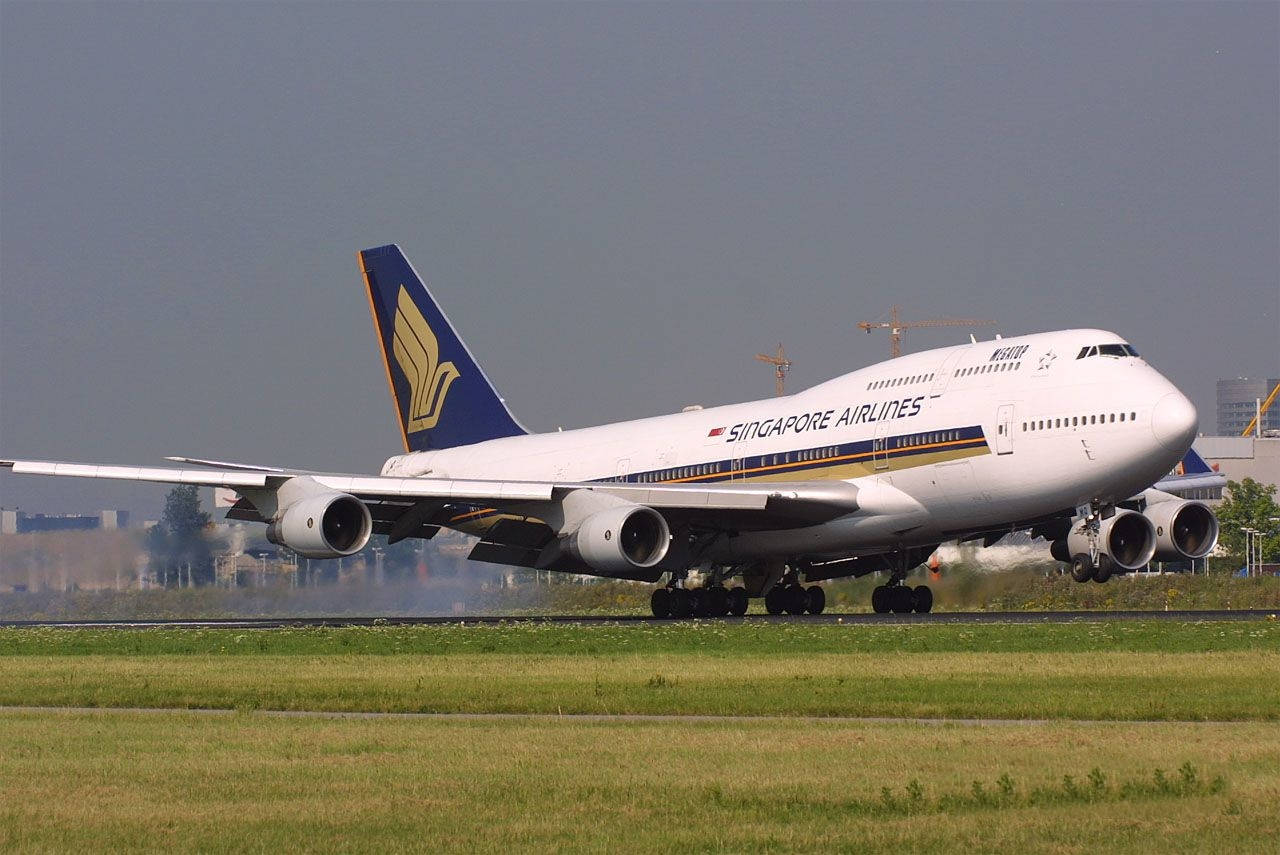 Singapore 桌布 壁紙 — 一架新加坡航空波音747在跑道上滑行，跑道上有綠色草地和遠處的機場建築，天空朦朧。