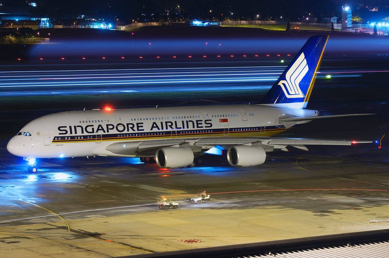 飛機 桌布 壁紙 — 一架新加坡航空的空中巴士A380在夜間滑過濕滑的停機坪，被鮮豔的藍色跑道燈光和機場光芒照亮。