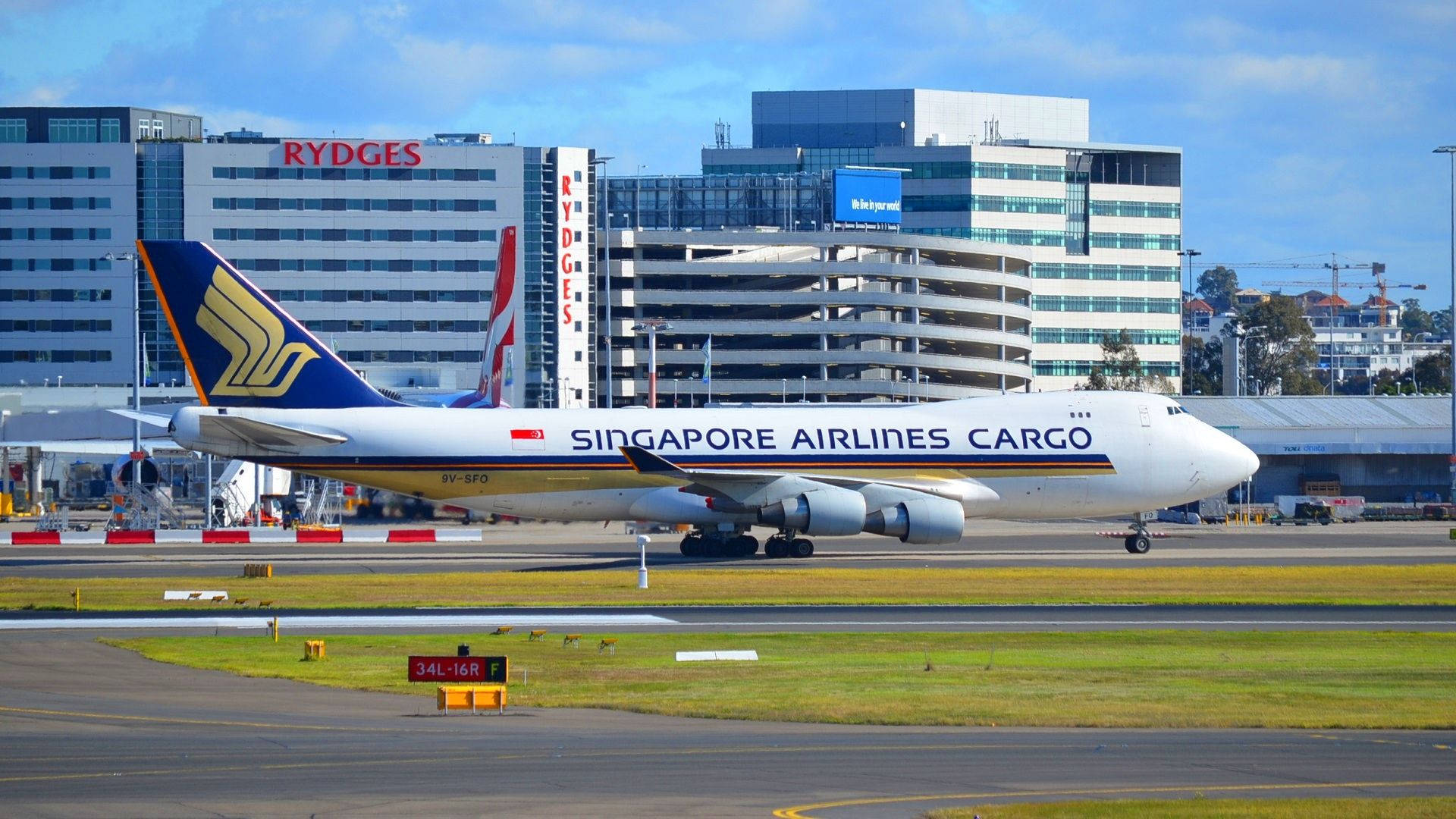 Buildings 桌布 壁紙 — 一架新加坡航空貨運的波音747在陽光明媚的機場停機坪上滑行，背景是現代建築和清澈的藍天。