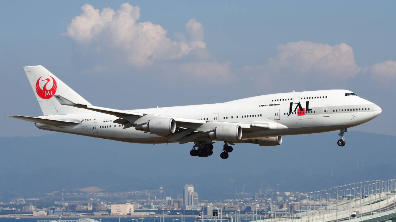 日本 桌布 壁紙 — 一架日本航空的波音747正在下降，起落架已放下，背景是藍天、蓬鬆的雲朵和遠處的城市景觀。